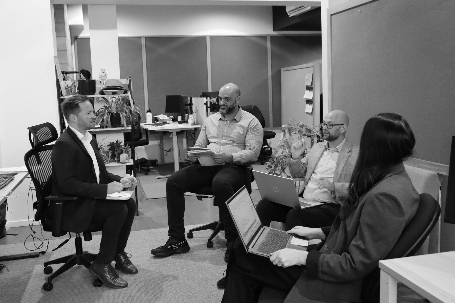 People in a meeting room sit in a circle, discussing with laptops and notepads.