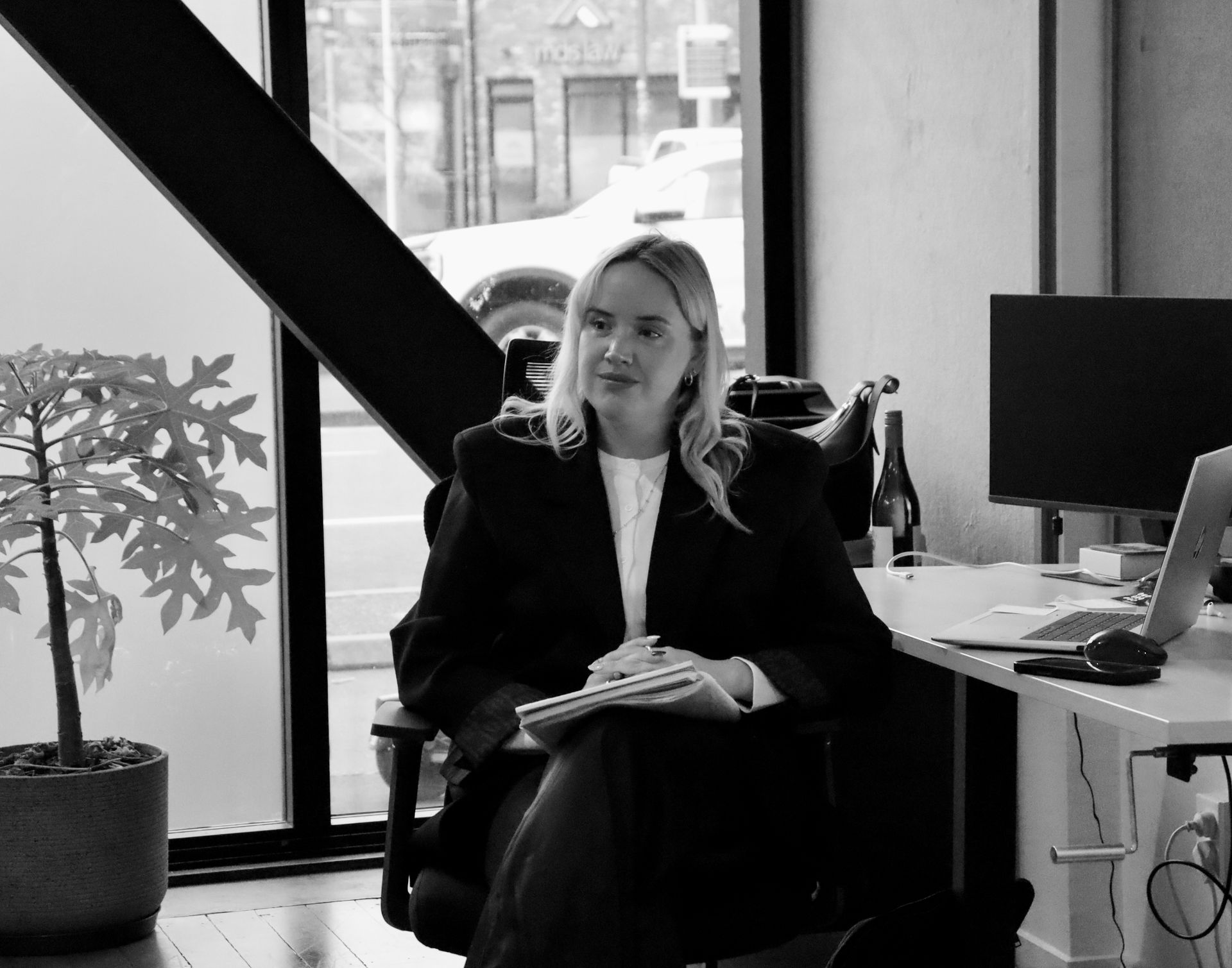 Woman seated at an office desk, holding papers with a computer and window behind her.