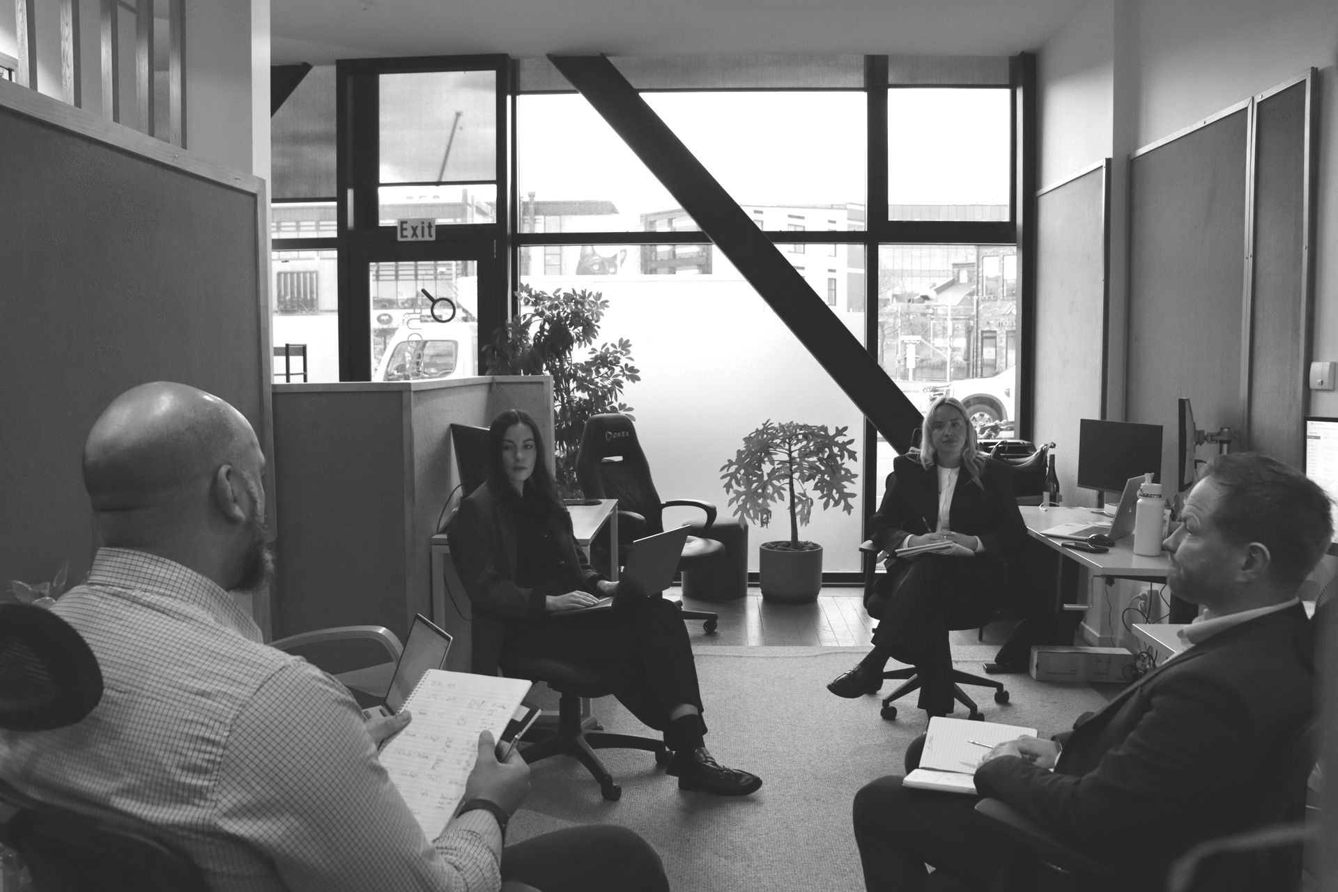 Four people sit in a modern office meeting space with large windows and plants.