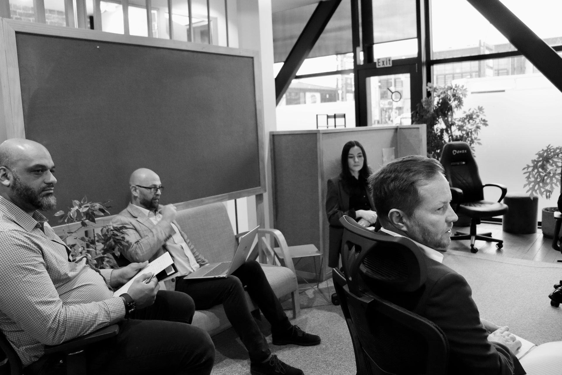 People are seated in a meeting room, listening and working with papers and laptops.