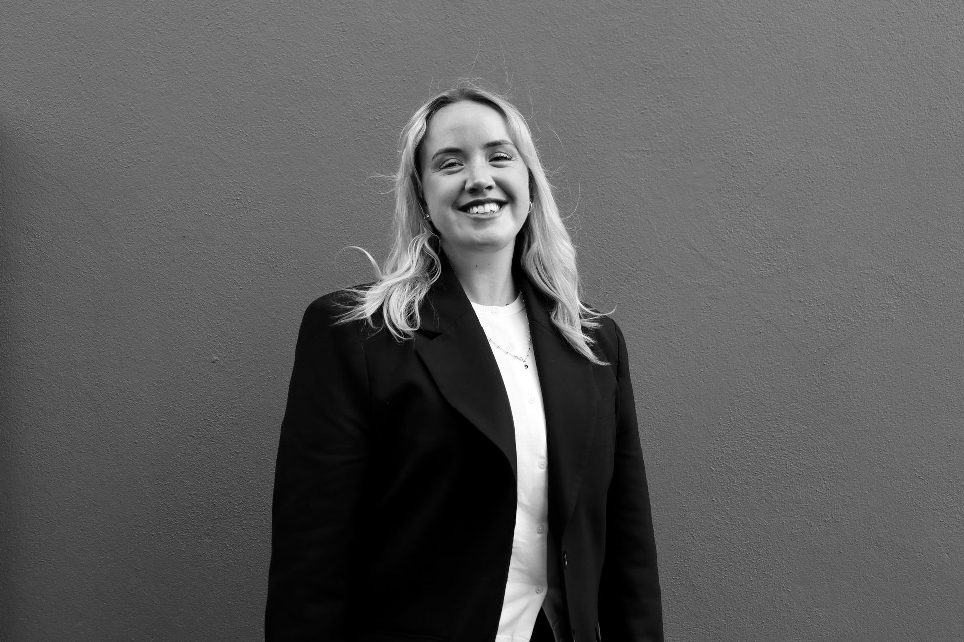 Smiling woman in a dark blazer standing against a plain wall