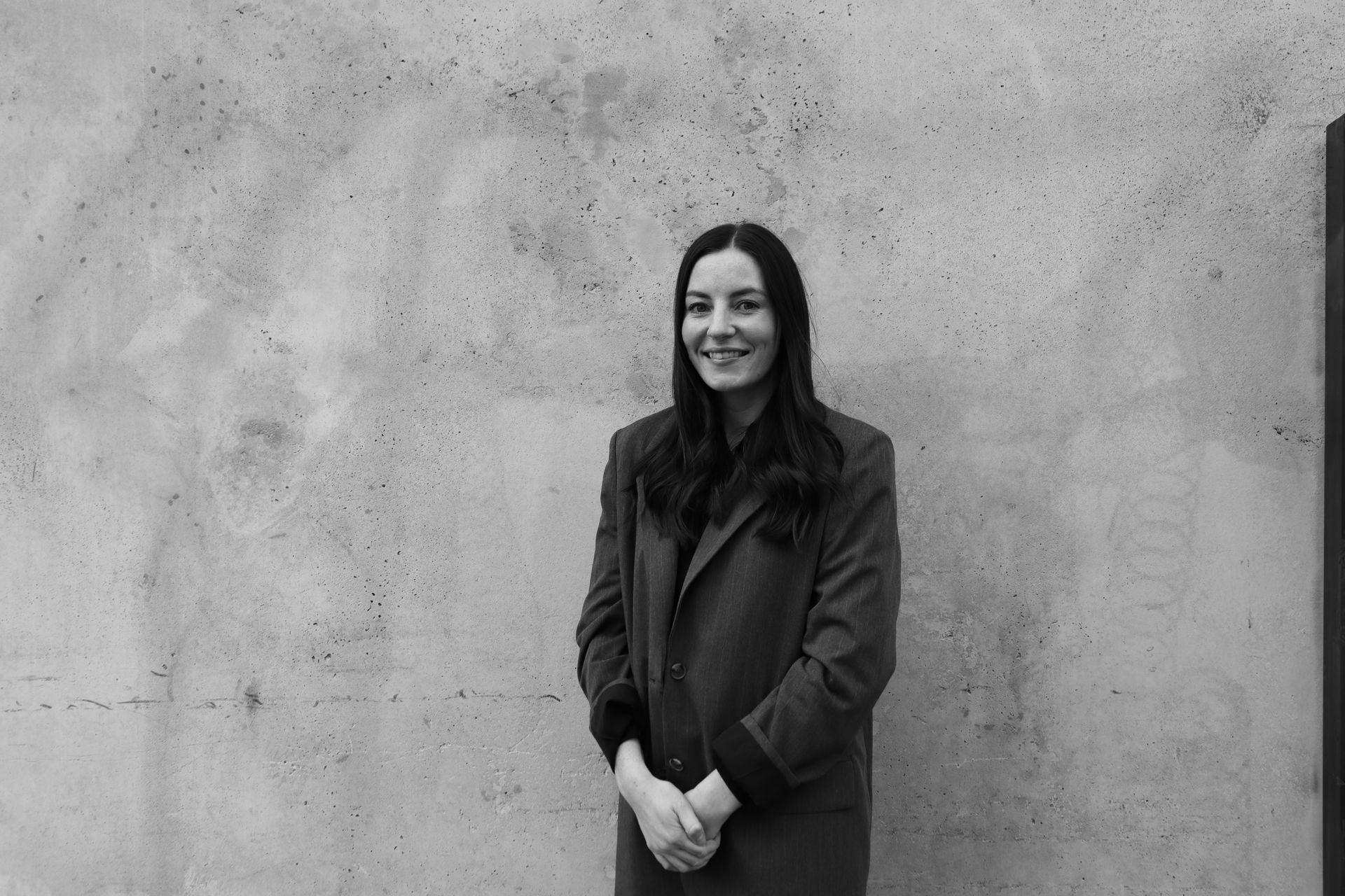 Woman in a dark coat standing and smiling against a plain concrete wall.