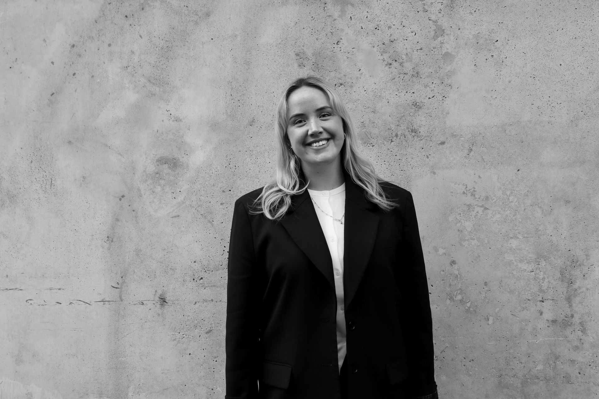 Smiling person in a dark blazer stands against a textured concrete wall.