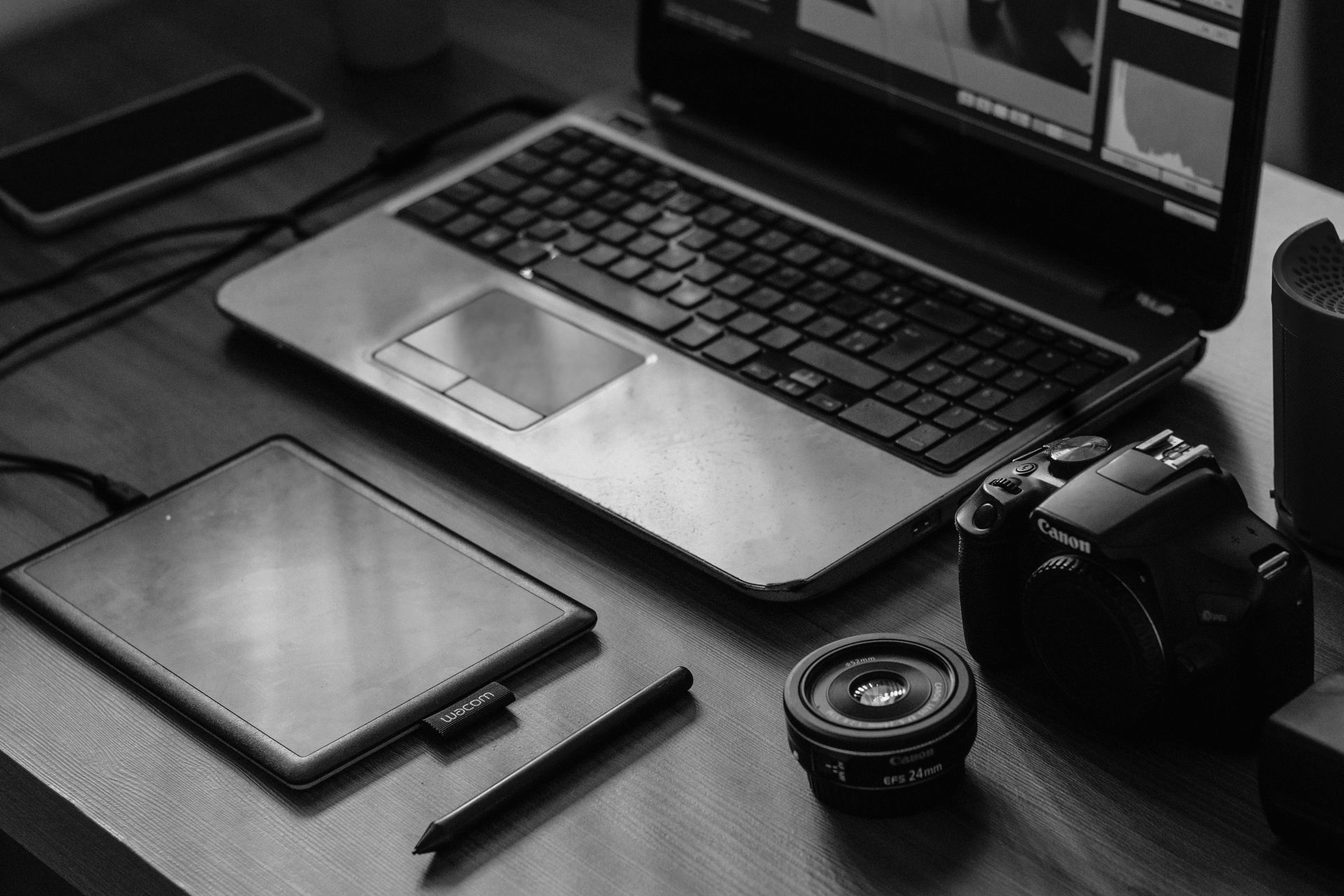 Black-and-white desk with laptop, tablet, camera, lens, and smartphone arranged on a workspace