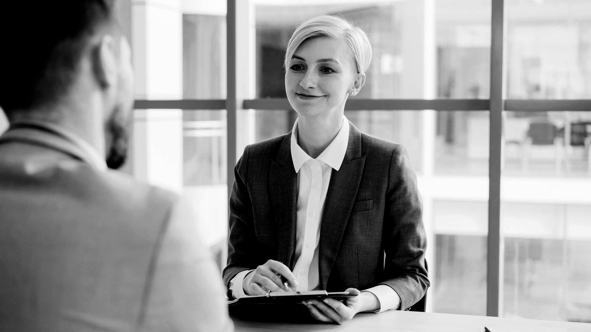 Two people in a business meeting, with a smiling woman seated across from another person near a window.