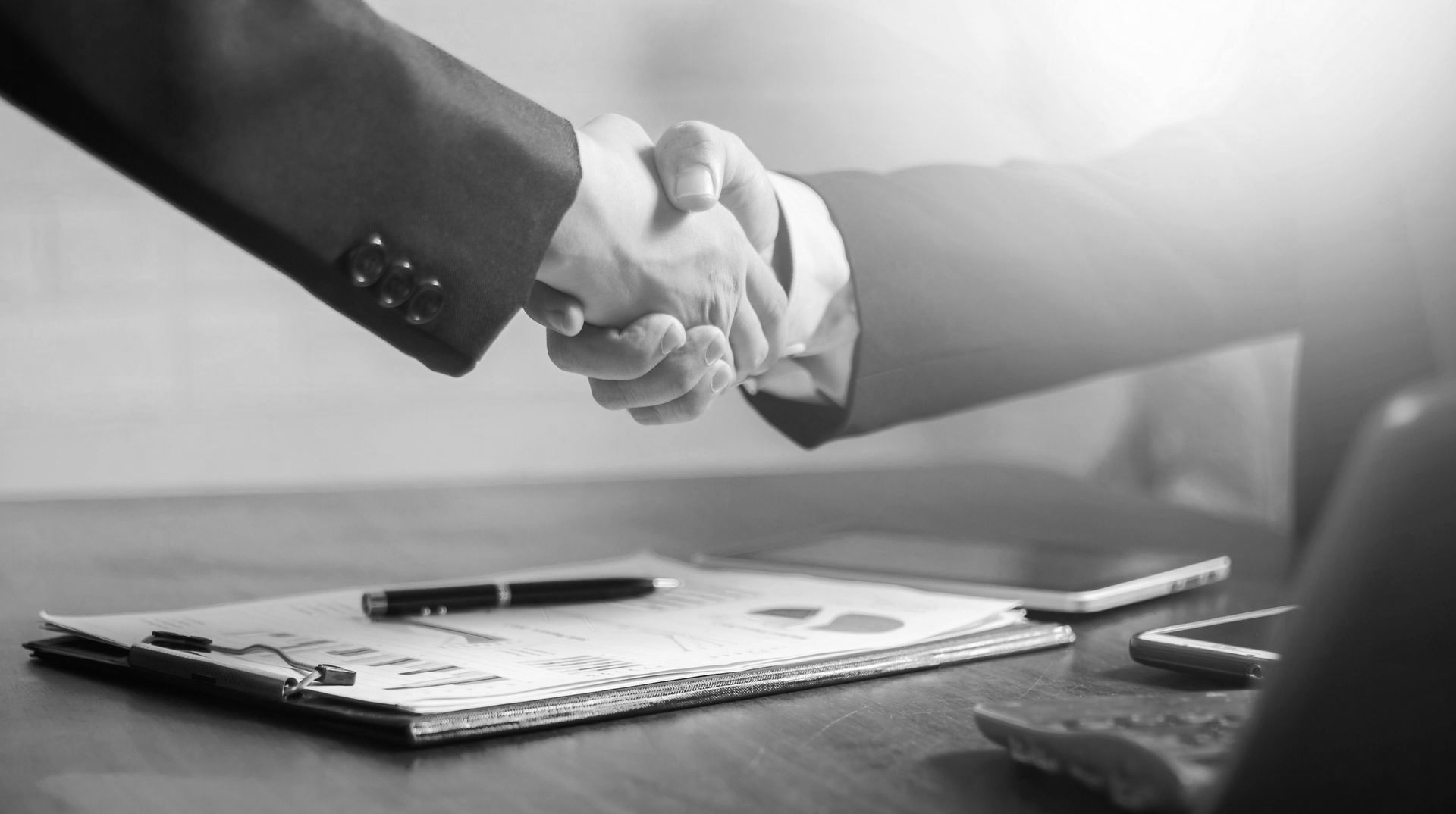 Two people shaking hands over a desk with documents and pens in black and white.