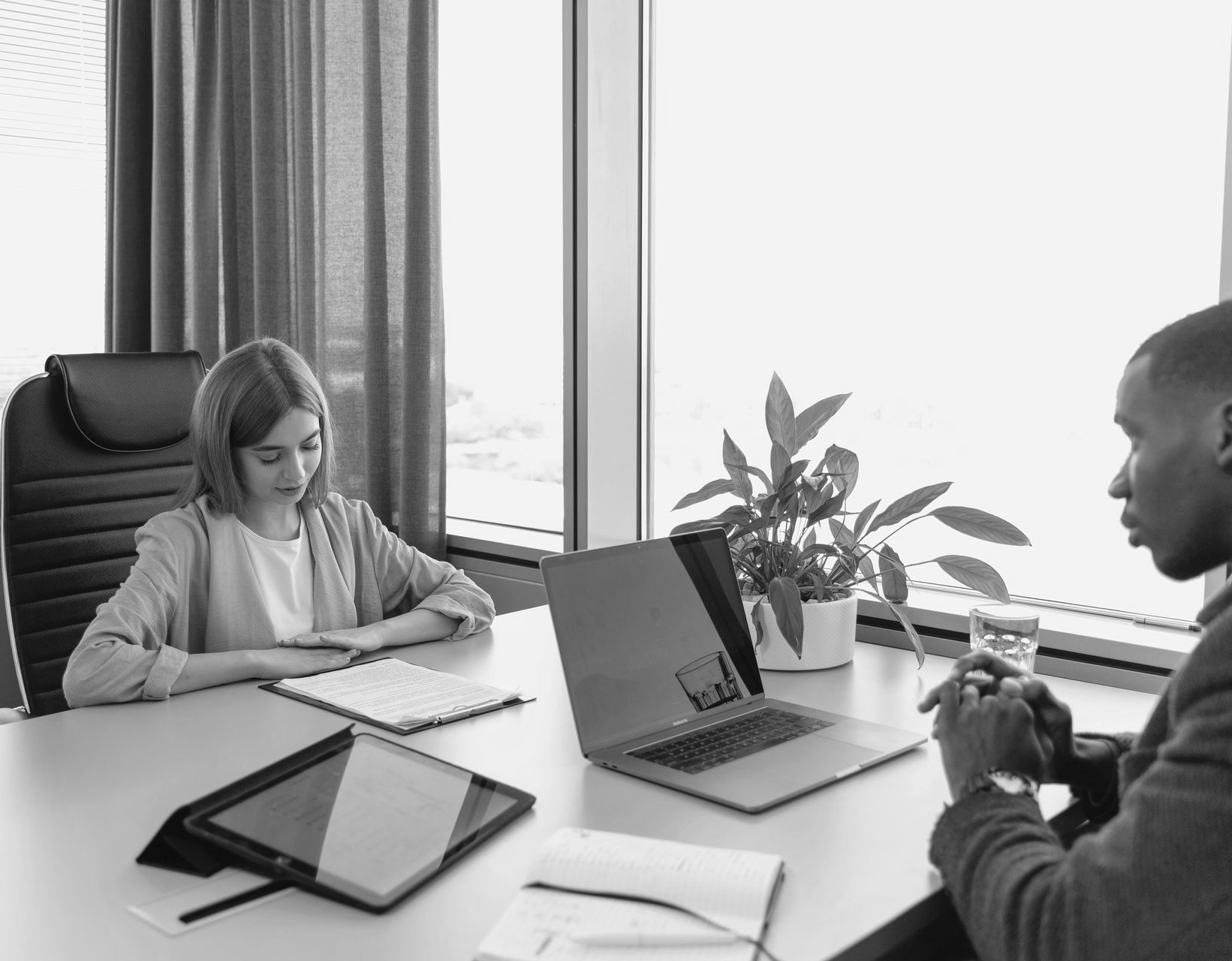 Two people meet at a desk with laptops and papers in a bright office.