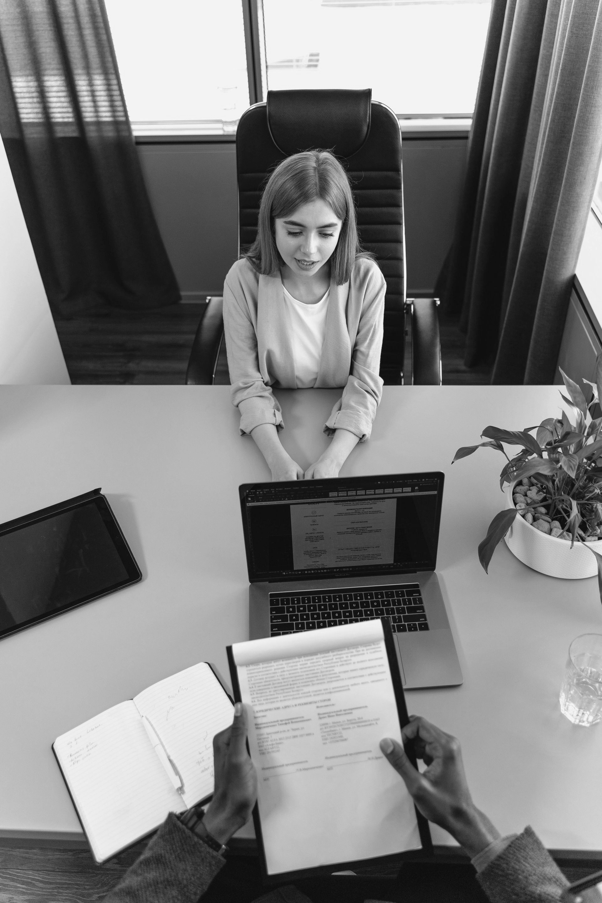 Person in an office meeting across a desk, reviewing documents on a laptop with notebooks and a plant nearby.