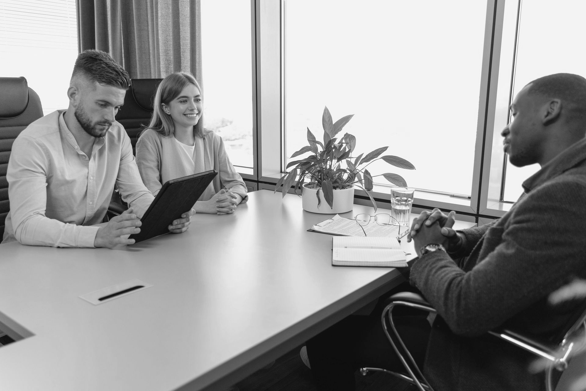 Three people in a meeting room discussing a tablet across a conference table.