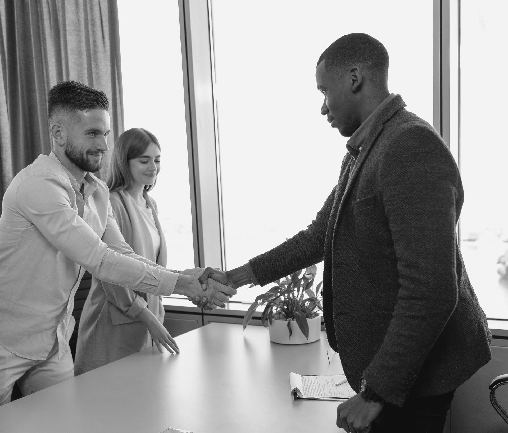 Two colleagues shaking hands across a desk in a bright office while a third person stands nearby.