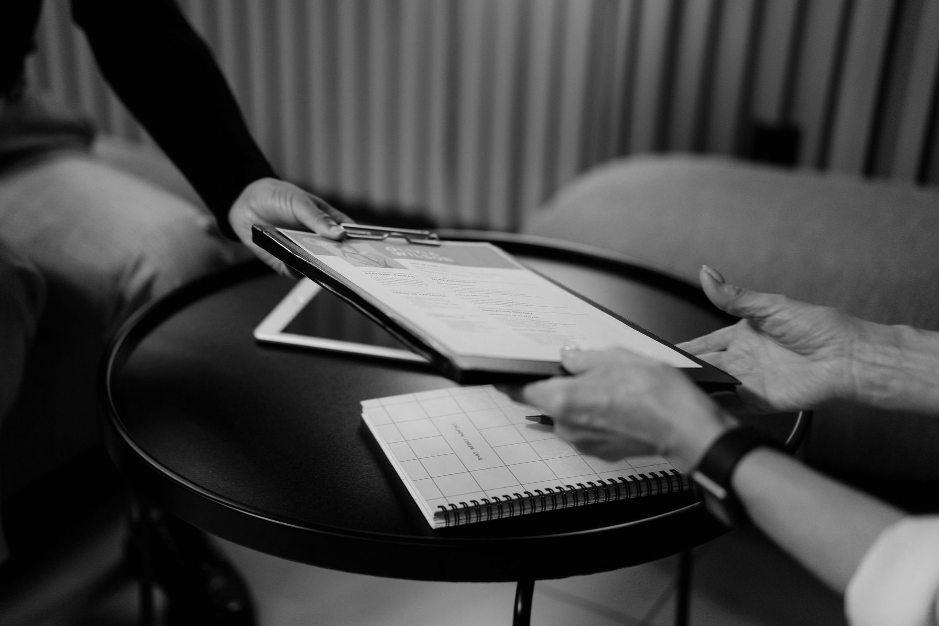 Hands holding a tablet and notebook over a small round table in a black-and-white setting