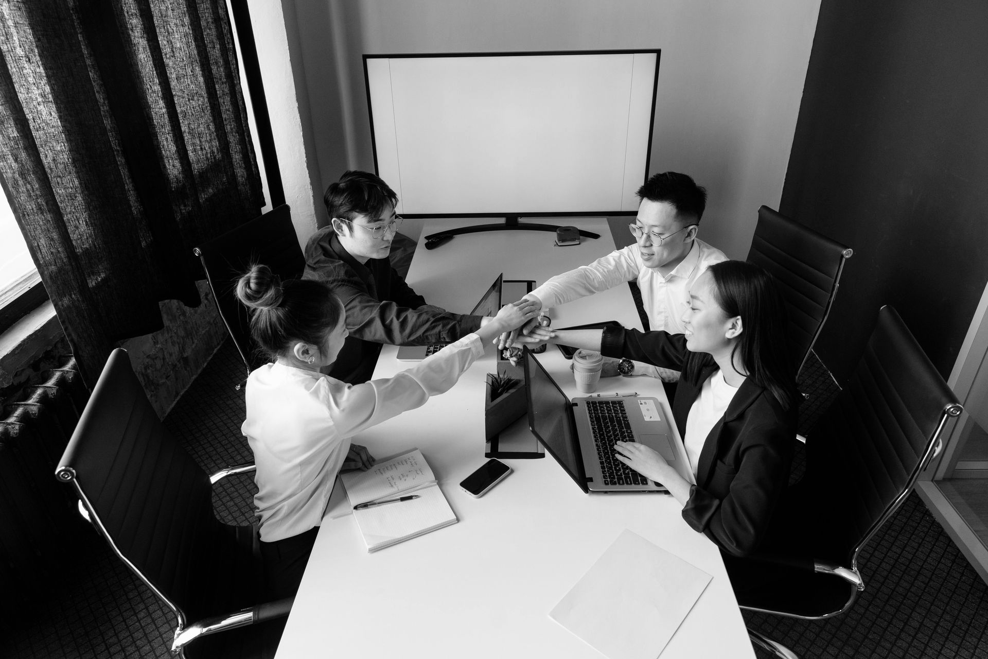 Four coworkers in a meeting room lean in and clink glasses around a conference table with laptops and notebooks.