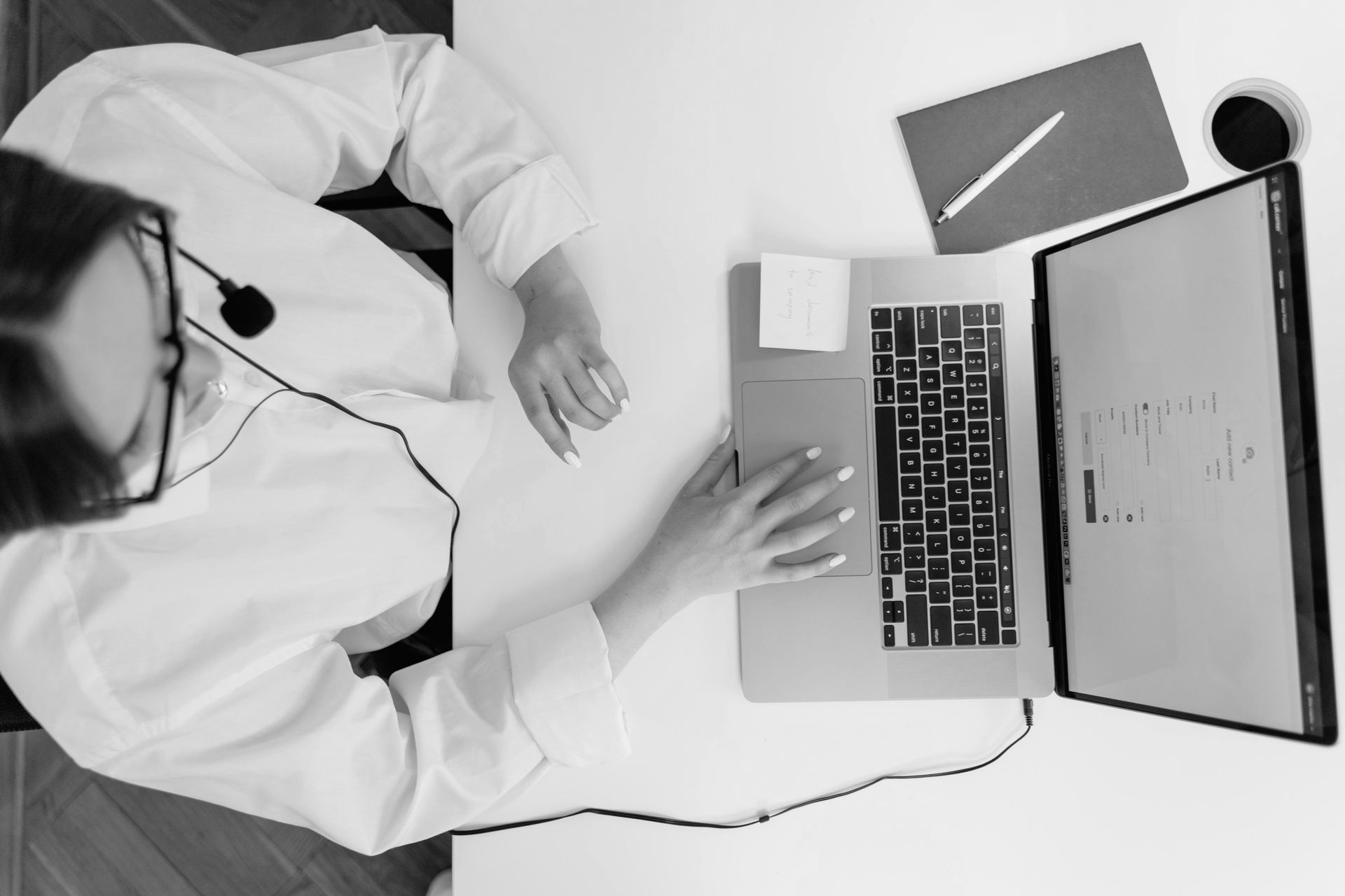 Person in white coat working at a laptop with headset, viewed from above on a desk with notebook and cup