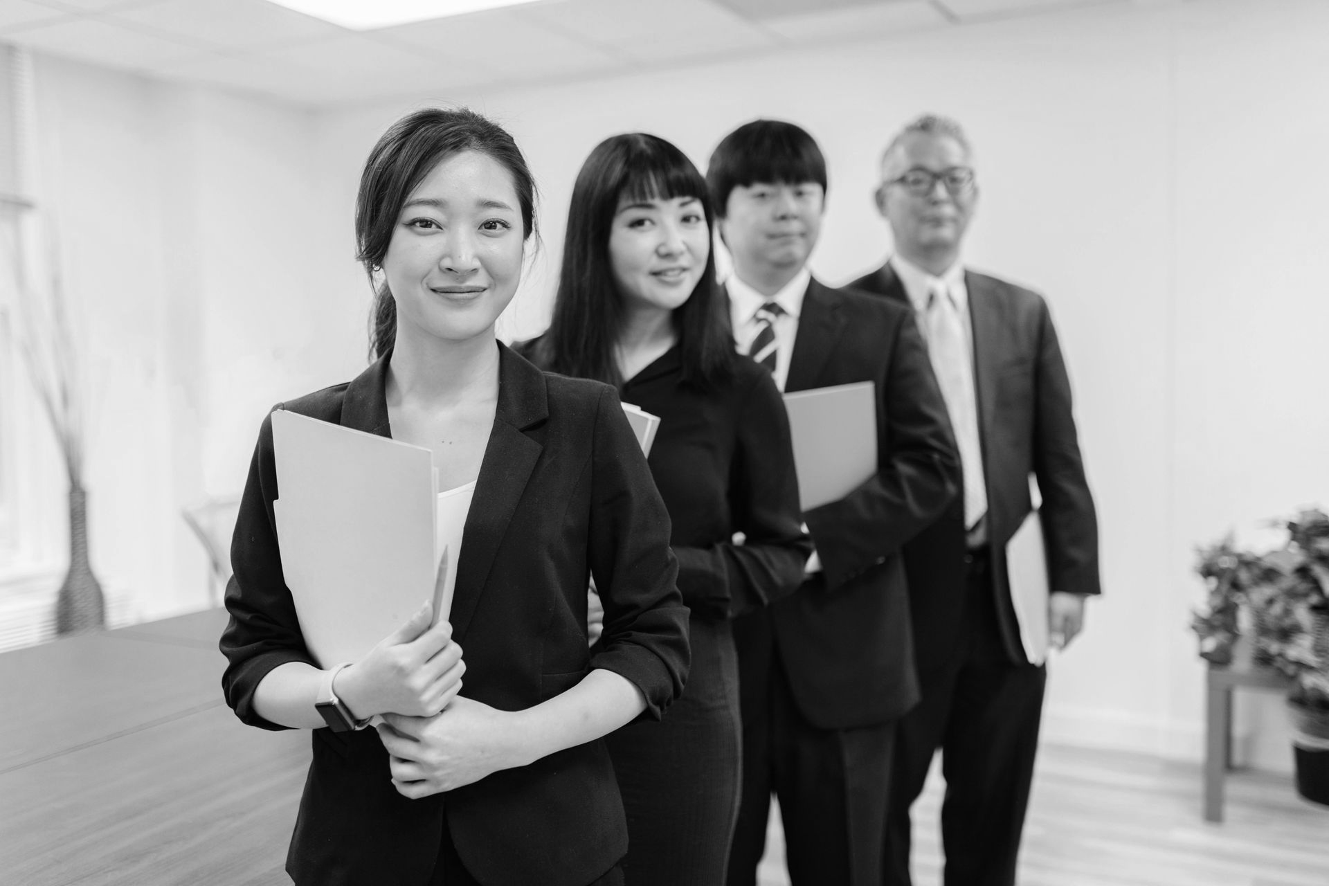 Four business professionals stand in a bright office hallway, holding documents and facing the camera.