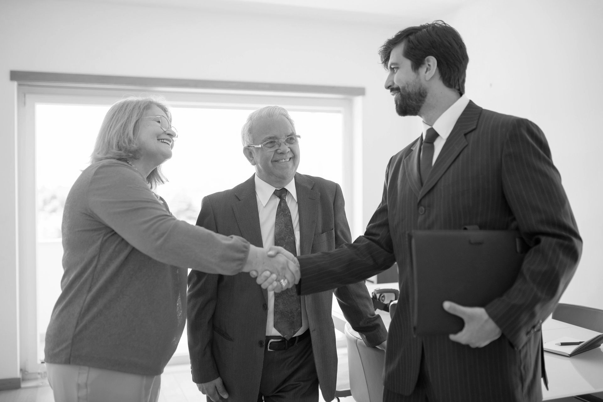 Three people in business attire shake hands in a bright office.