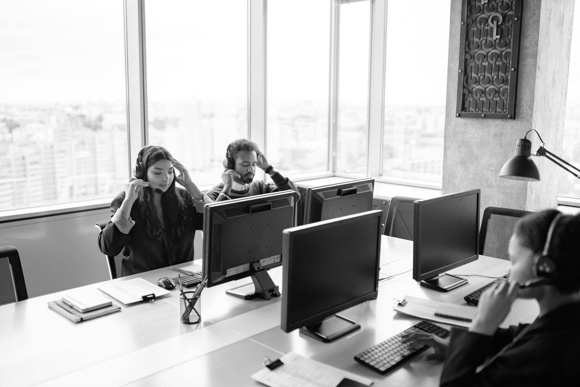Office meeting with three people around a desk and computer monitors near large windows.