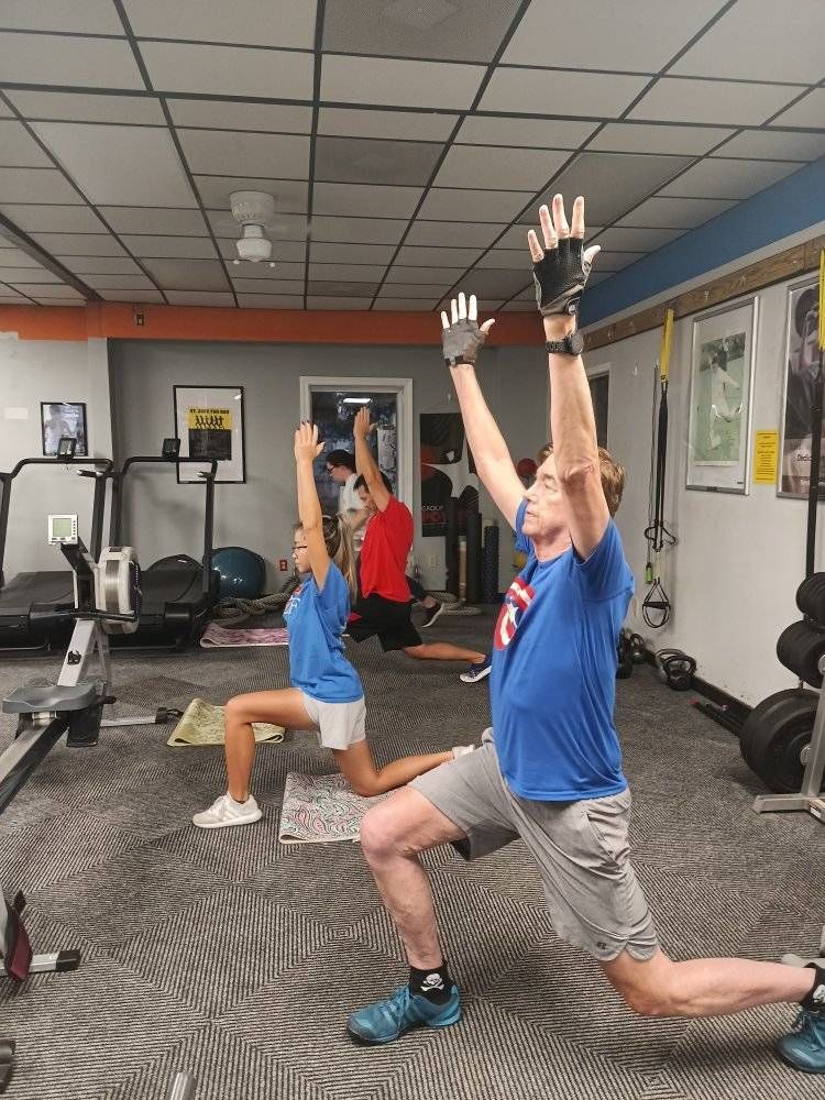 People in gym lunging with arms raised. Indoors with exercise equipment.