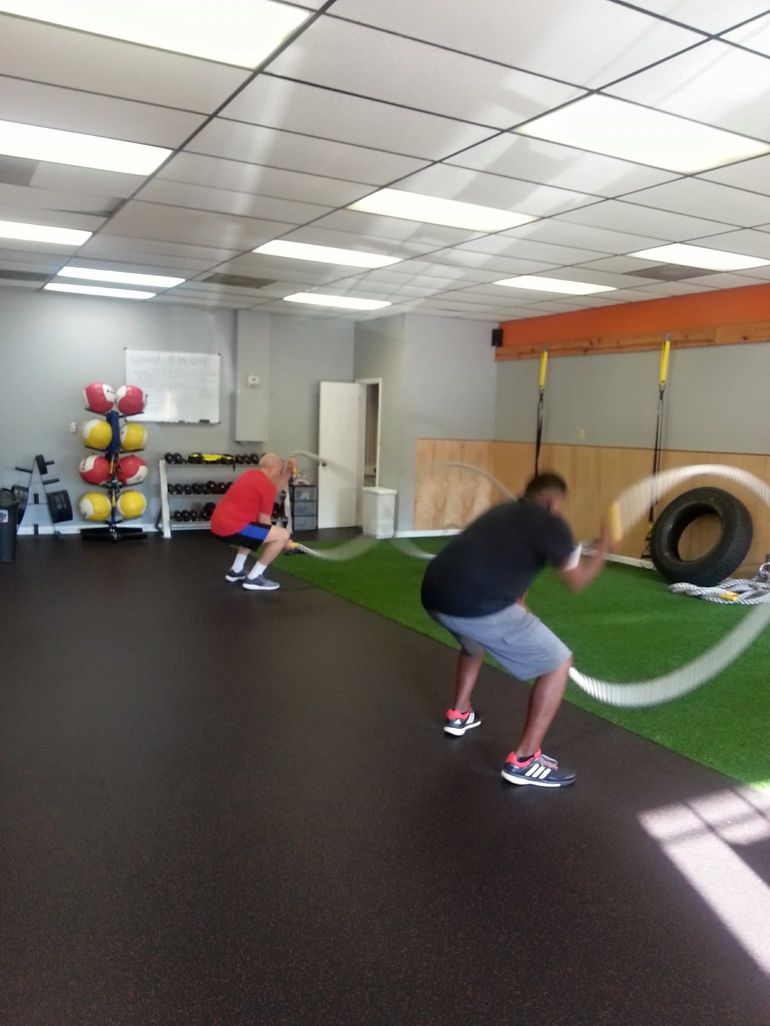 Two men working out with ropes in a gym. One wears red, the other black, both in athletic shoes.