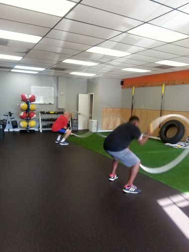 Two men working out with ropes in a gym. One wears red, the other black, both in athletic shoes.