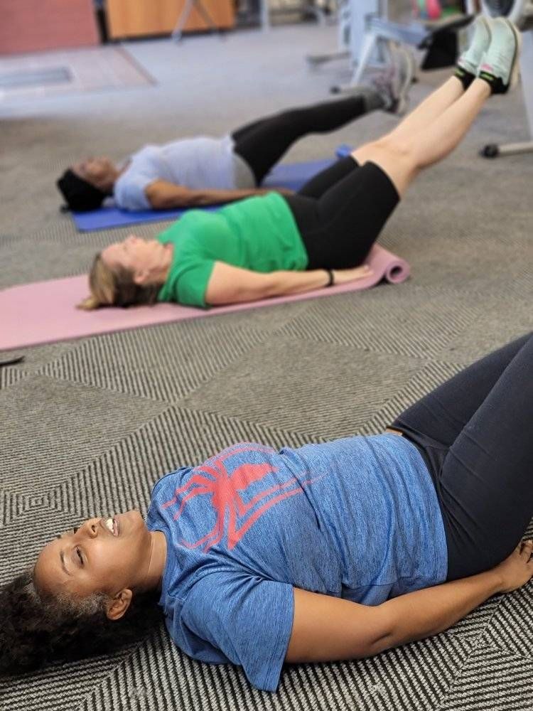 Three women exercising, lying on mats with legs raised, in a gym setting.
