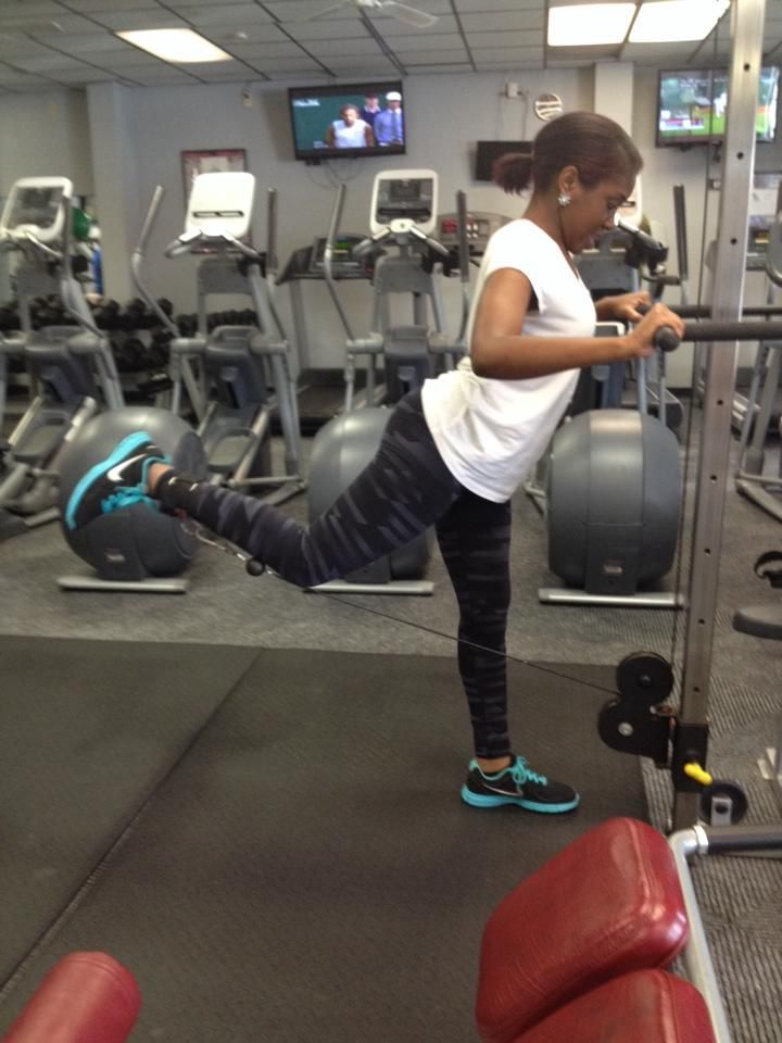 Woman in gym doing cable kickback exercise. She wears a white shirt and black leggings, using a cable machine.