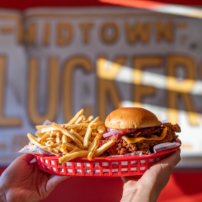 A  person holding up a basket of fries with Clucker's Cowboy Clucker Sandwich. In the background is a sign that says 