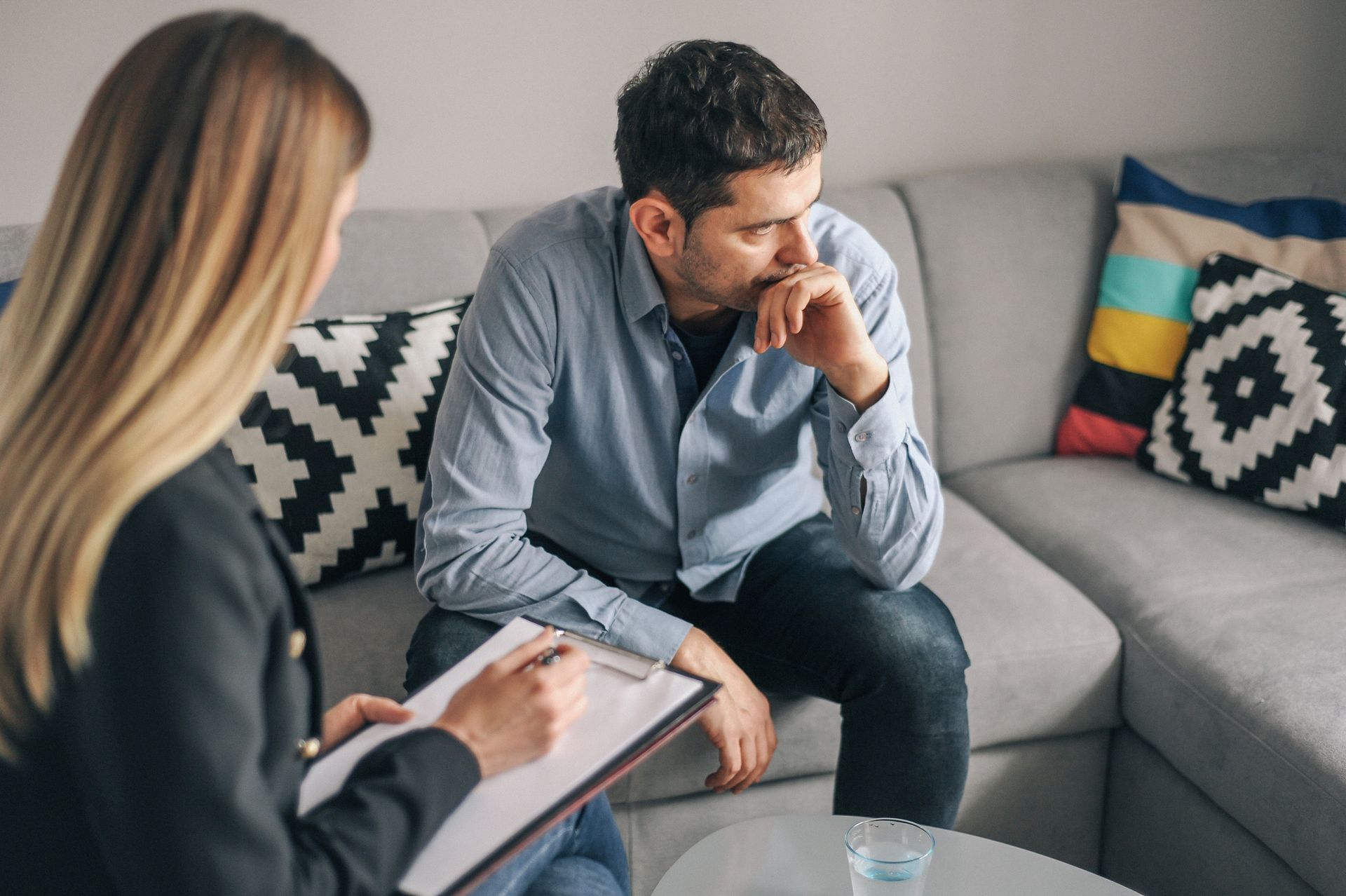 A man is sitting on a couch talking to a woman who is holding a clipboard.