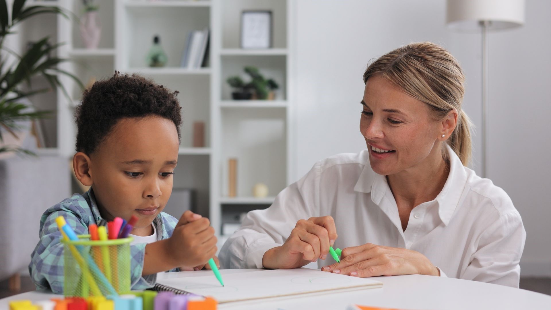 A woman is helping a young boy with his homework at a table.