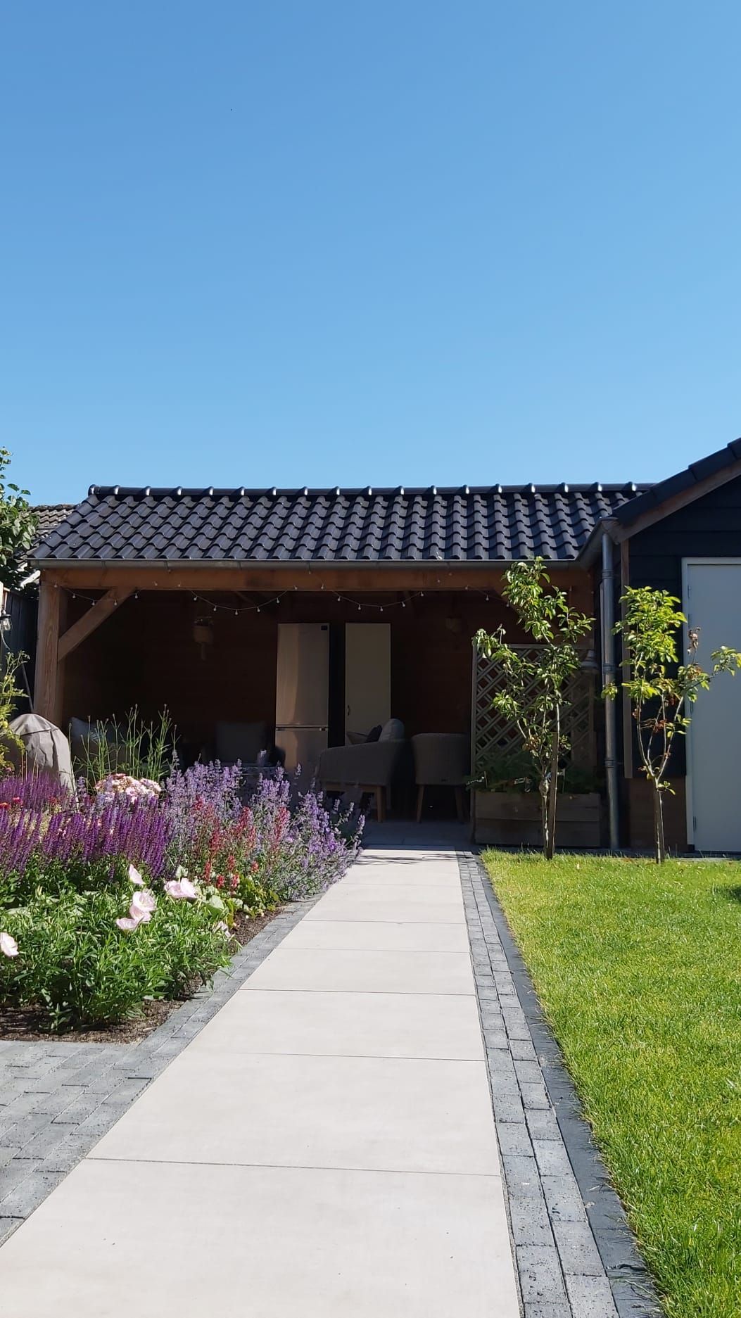 Achtertuin met pad van keramische tegels, gazon en terras met bloeiende planten.