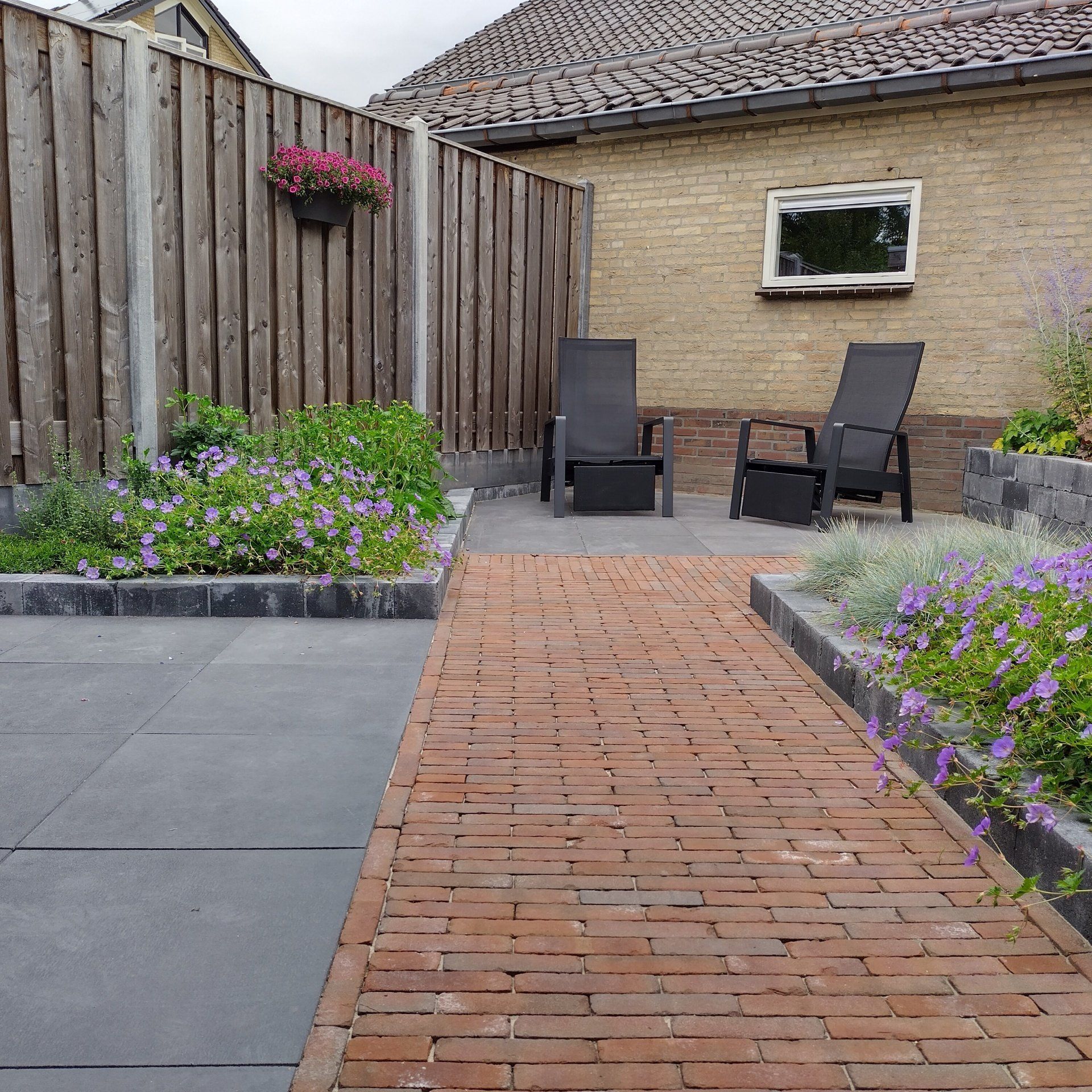 Een bakstenen pad leidt naar een terras met stoelen en bloemen