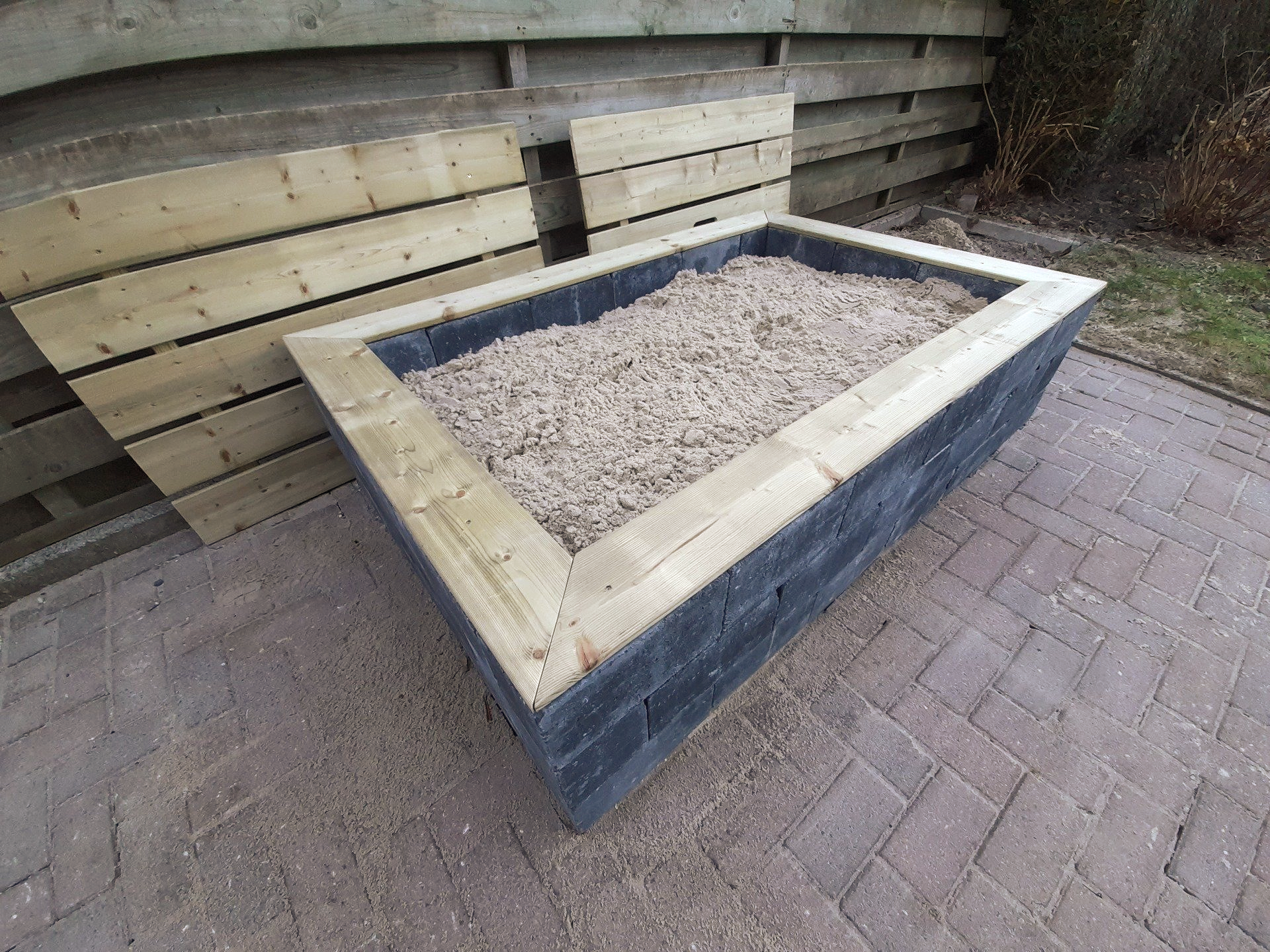 Een zandbak van hout en steen staat op een bakstenen terras. 