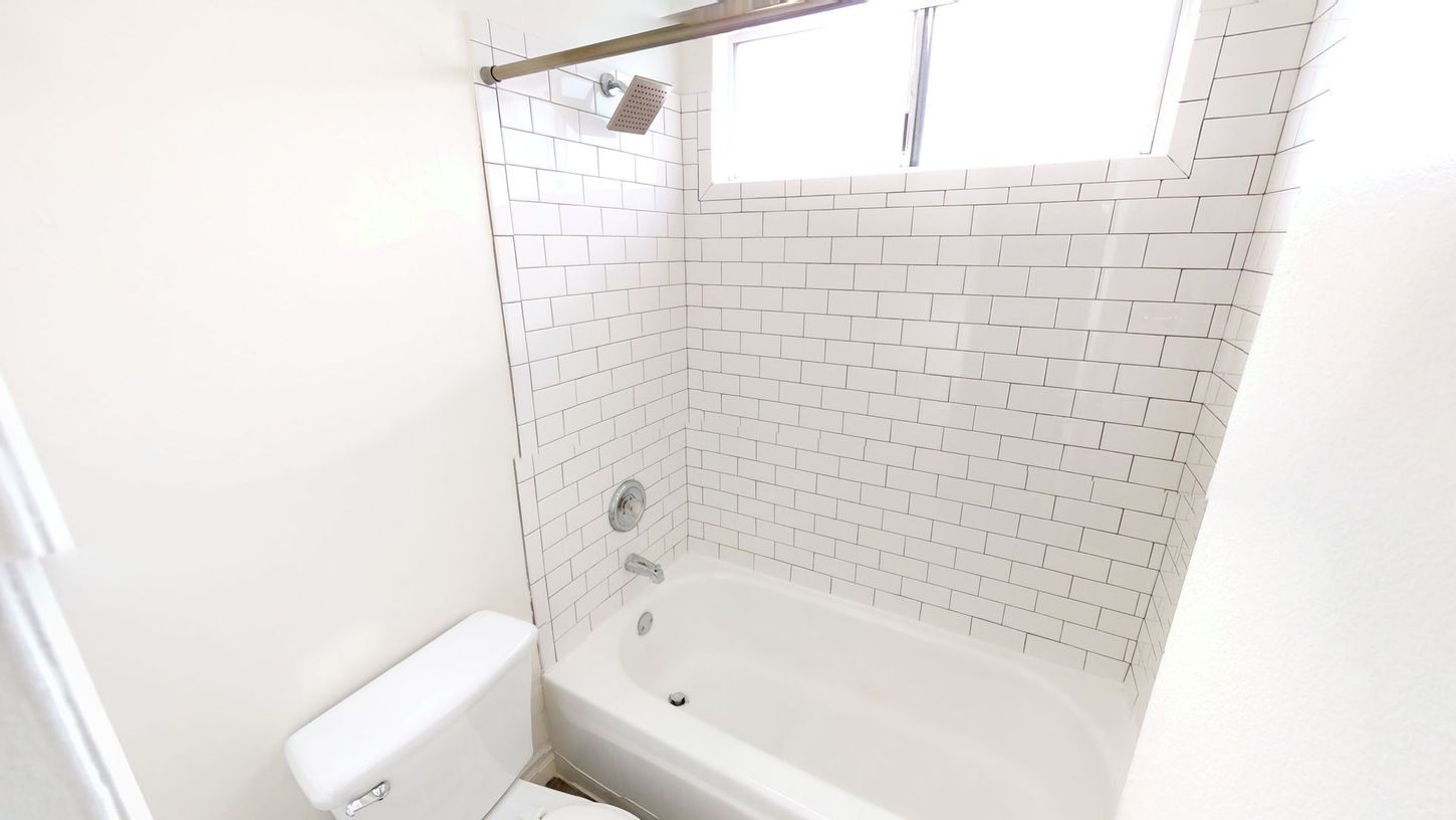 White bathroom with a tub, toilet, and tiled shower area, with a window.