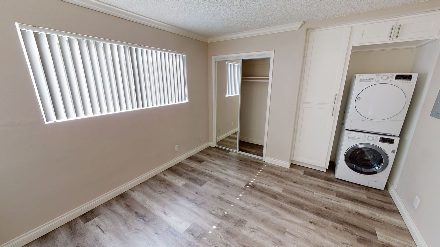 Empty room with wood-look floor, blinds, closet, stacked washer/dryer, and storage cabinets.