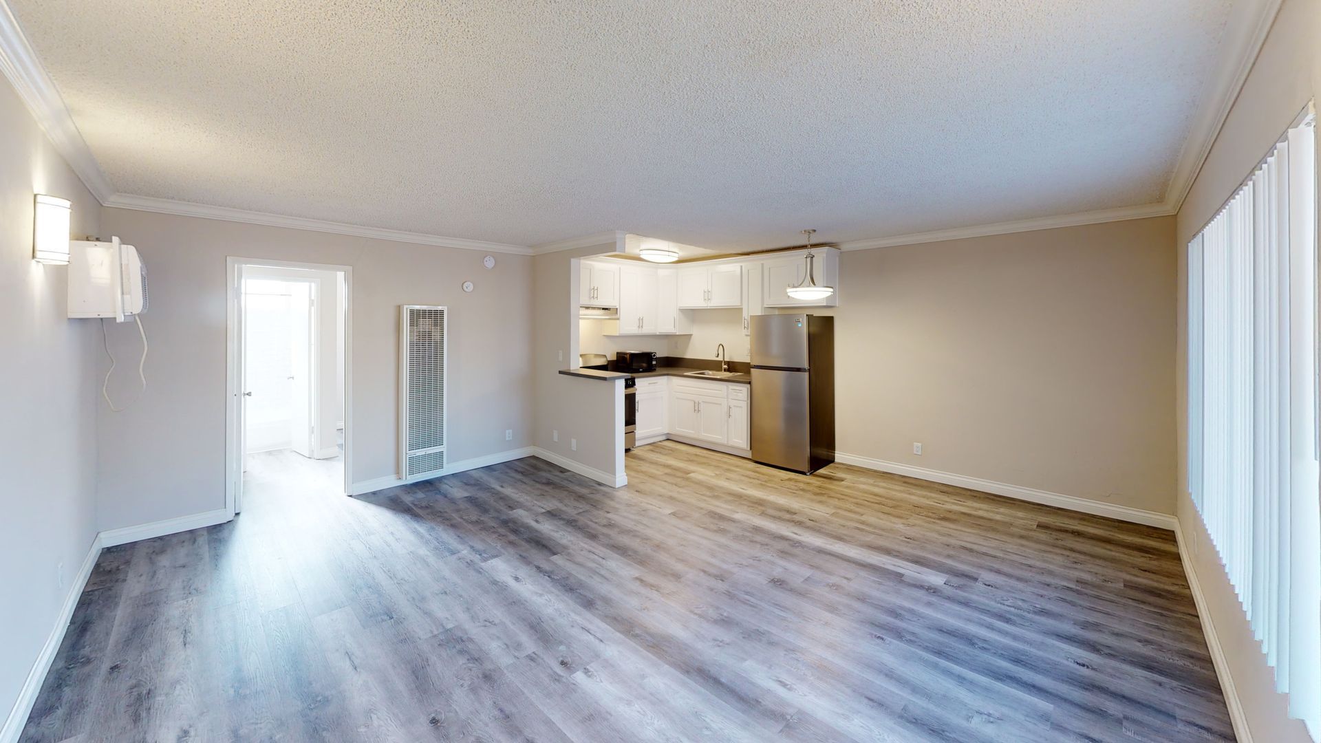An empty, open-concept apartment interior with grey wood-look flooring, a kitchen, and a bright window.