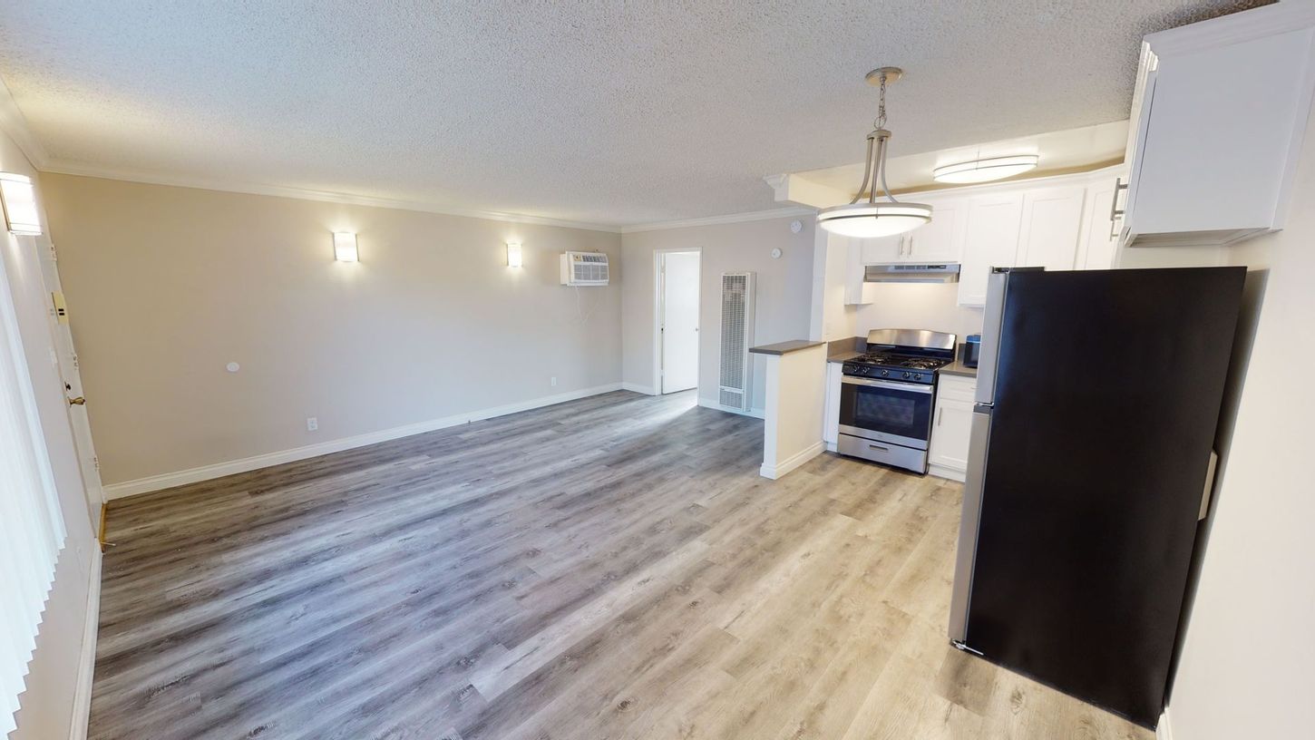 Empty apartment interior, light wood floors, kitchen with black fridge, white cabinets, and stainless steel appliances.