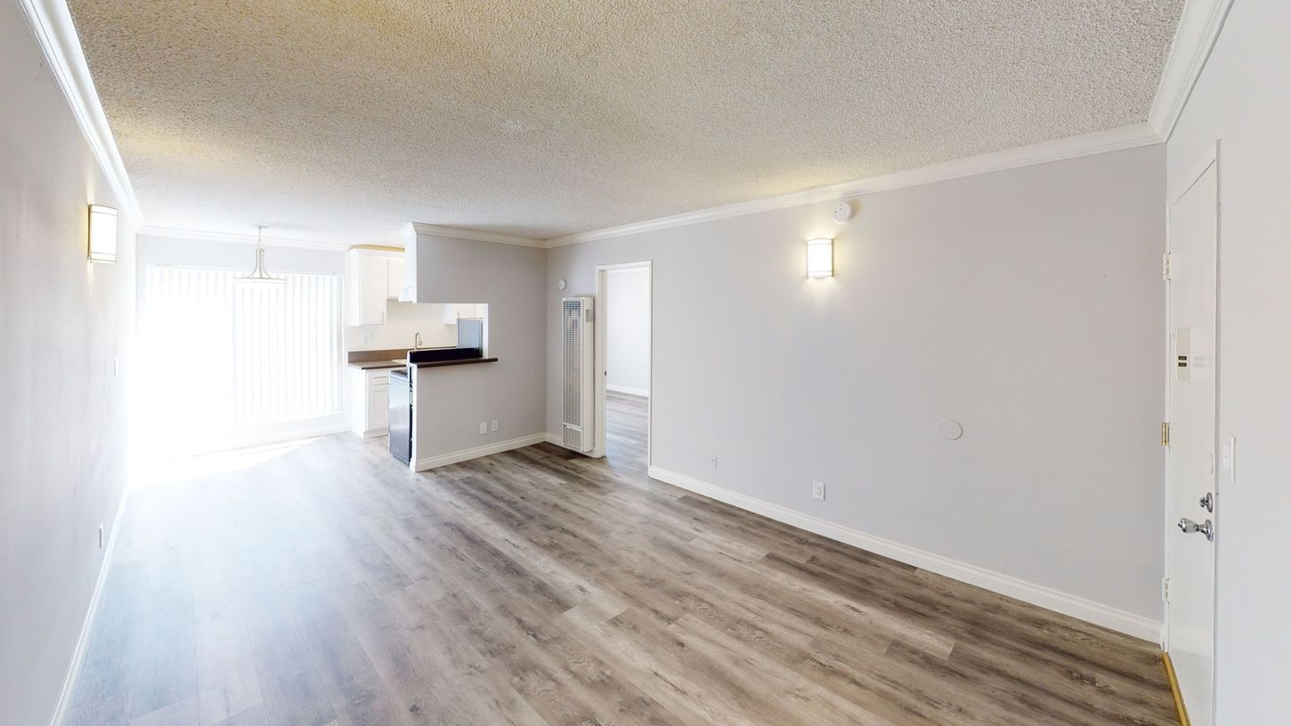 Empty living room with wood flooring, white walls, and a kitchen area.