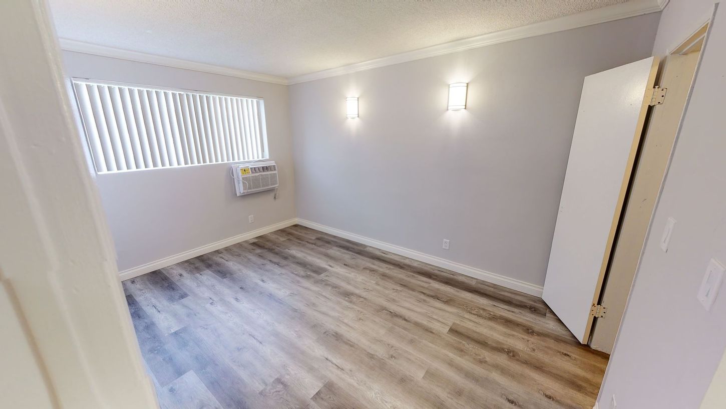 Empty bedroom with light gray walls, wood-look flooring, and a window with blinds.