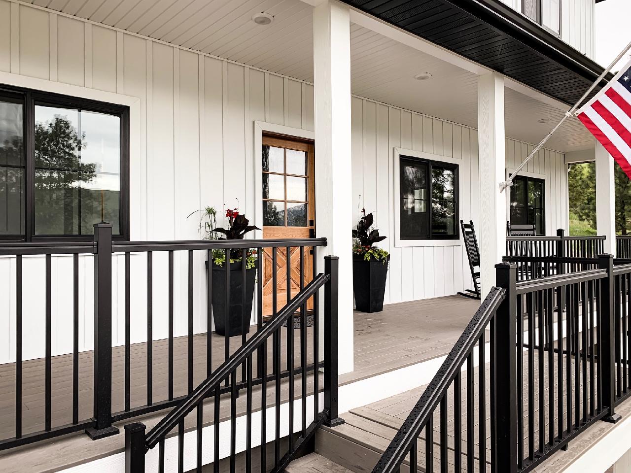 White farmhouse porch with black railing, windows, and door, featuring an American flag.
