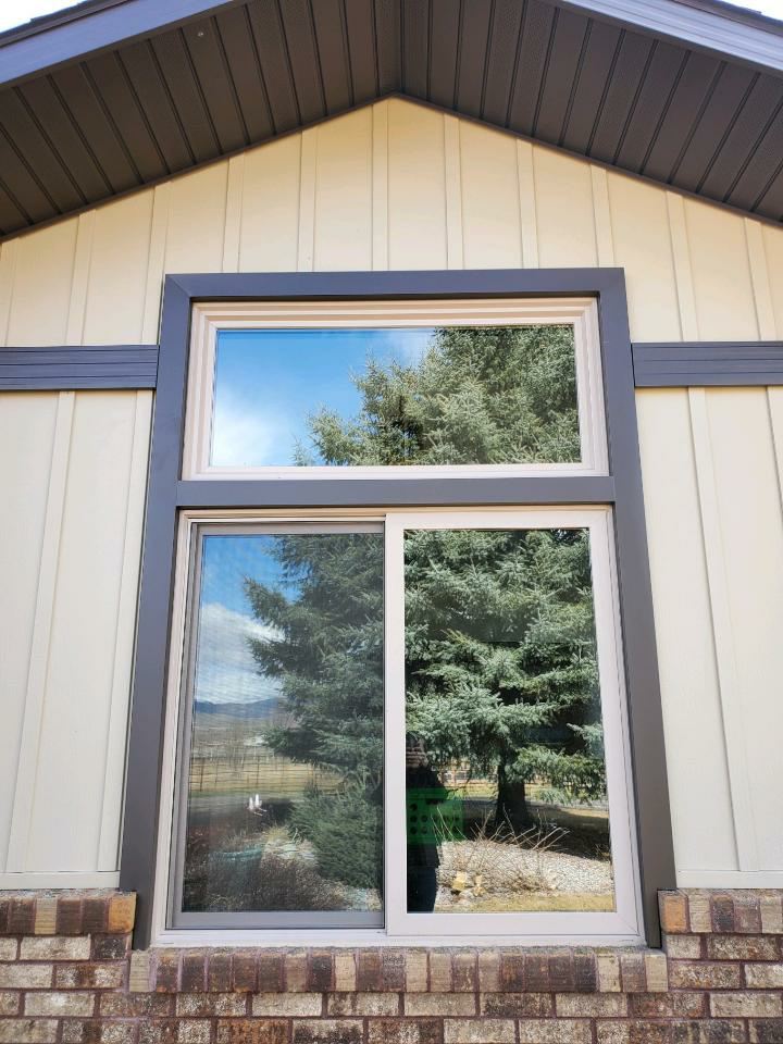 Window with a rectangular frame; top and bottom panes reflect a tree and sky. Beige siding and brick base.