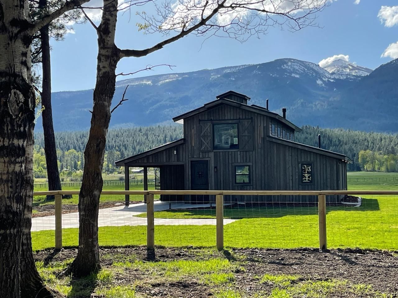 Rustic wooden barn with porch, fence, and mountain backdrop on a green field.