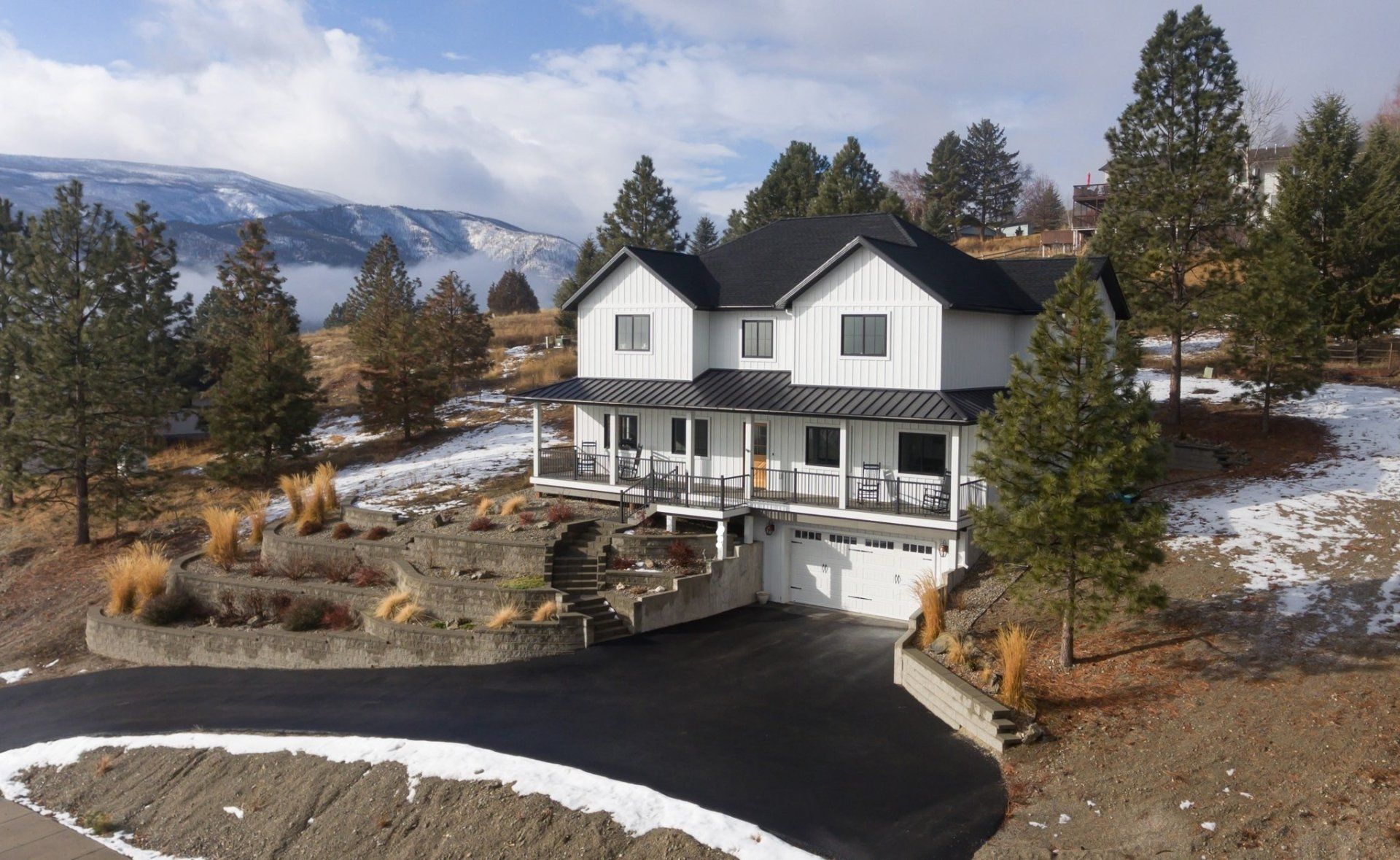 Two-story white house with black roof, on a hill with a driveway and snowy landscape.