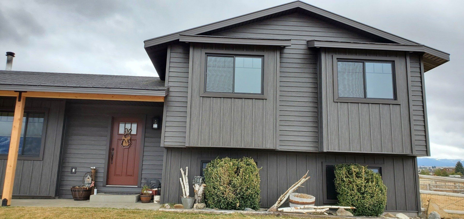 Gray house with red door and two second-story windows. Overcast sky and green bushes in front.