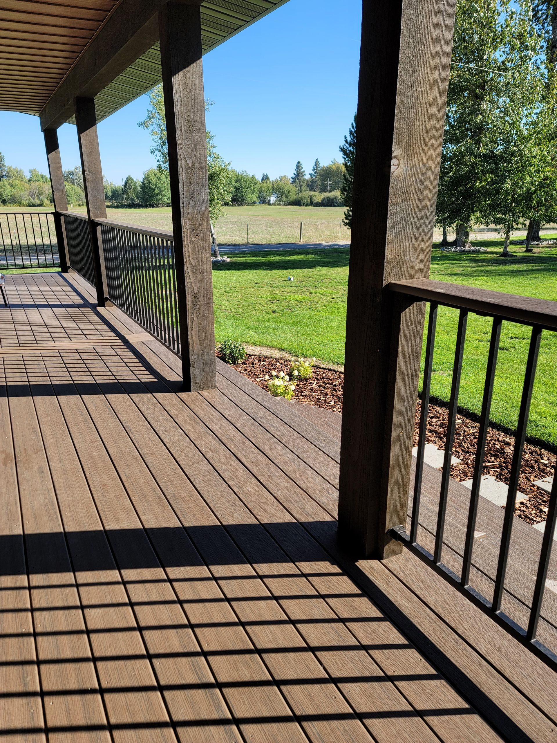 Wooden deck with railing and supports, overlooking green lawn and trees under a blue sky.