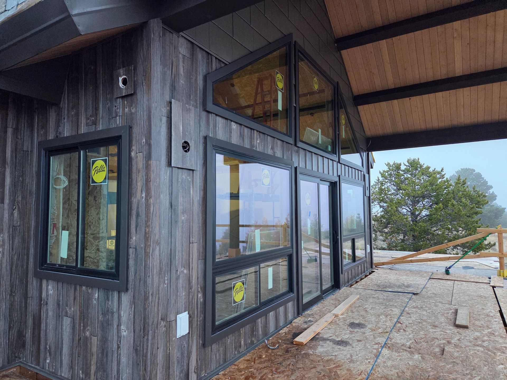 Exterior of a cabin under construction with gray wood siding and large black-framed windows overlooking a foggy landscape.