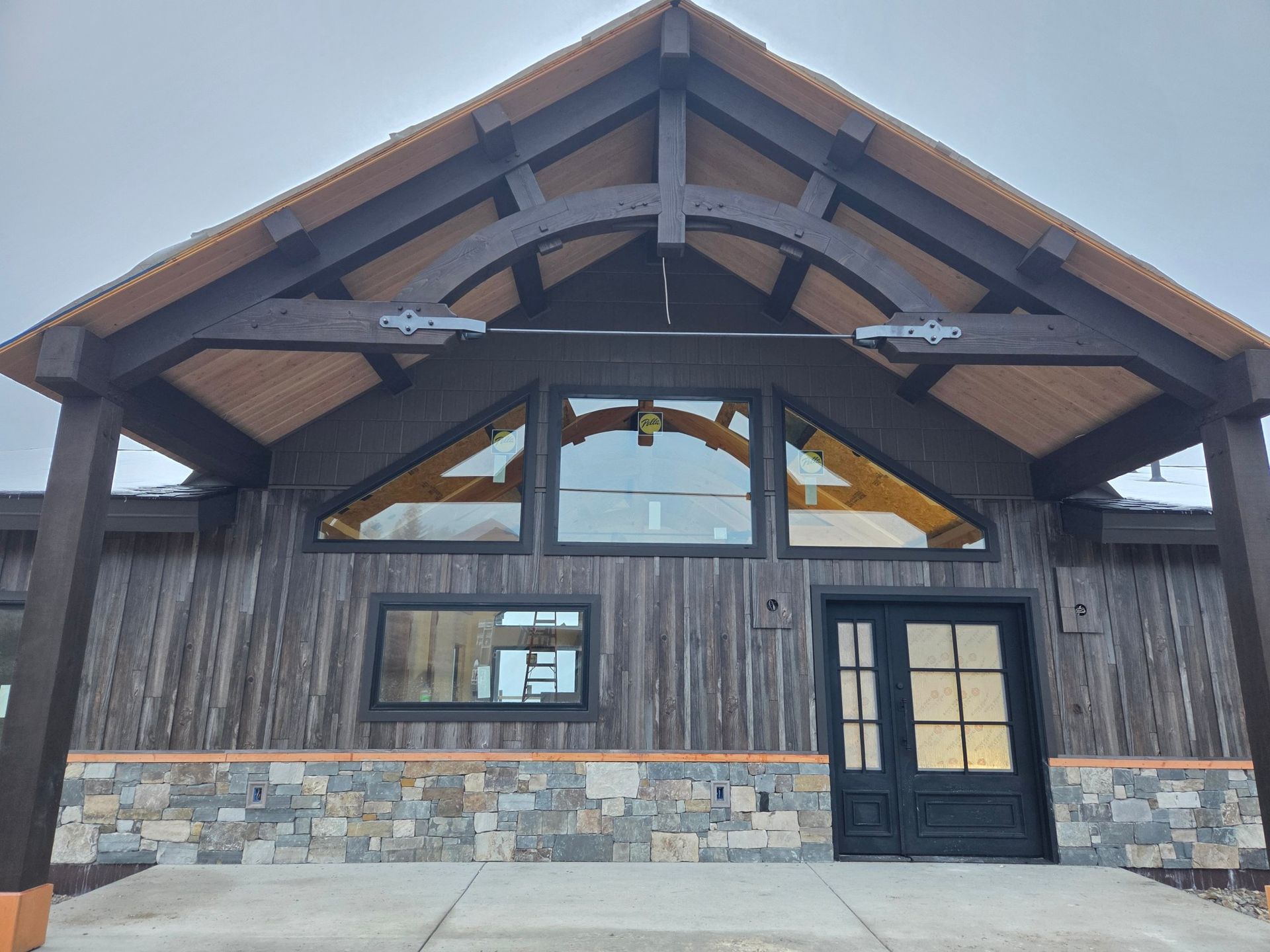 Rustic-style building exterior with wooden beams, stone accents, and glass windows. Cloudy sky overhead.