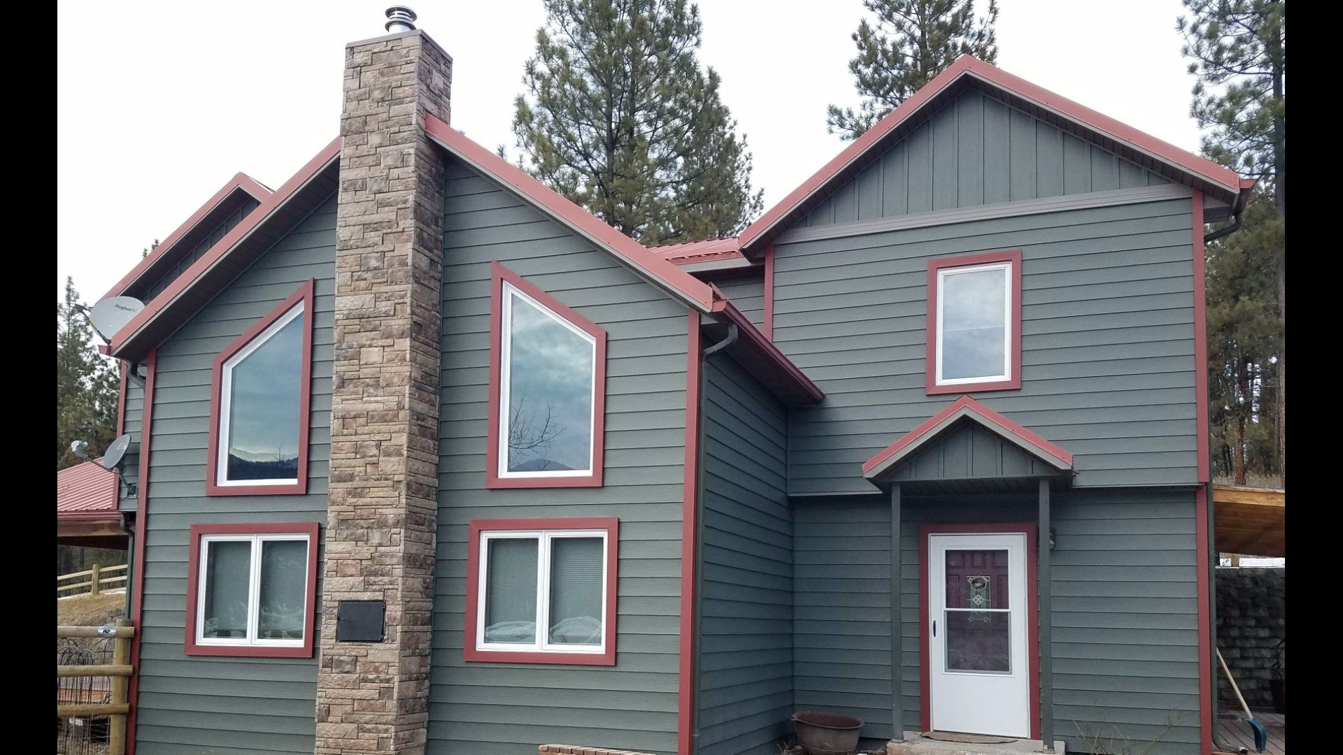 Two-story house with green siding, red trim, stone chimney, and multiple windows against a backdrop of trees.