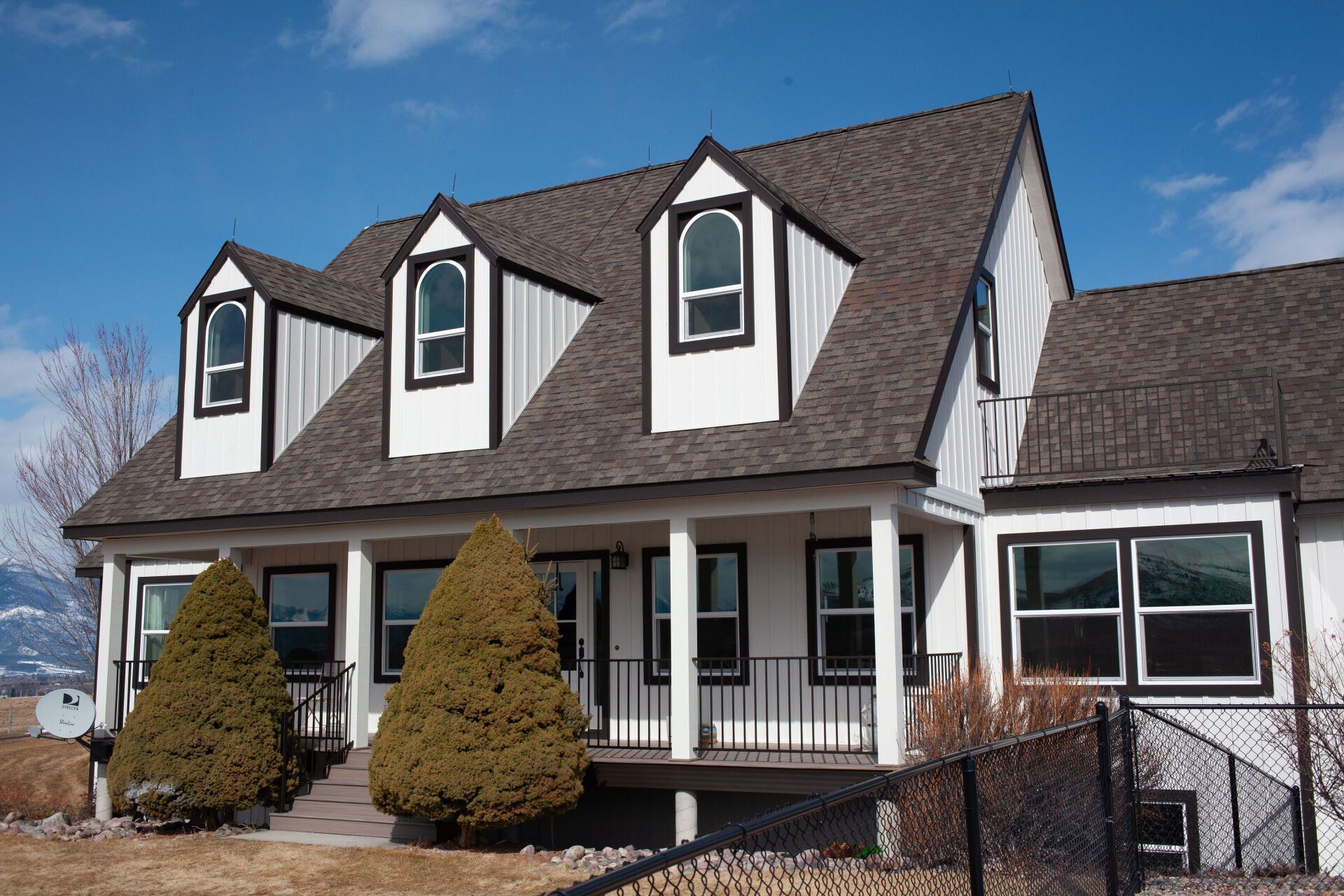 Two-story white house with black trim, three dormers, and a porch, set against a blue sky.
