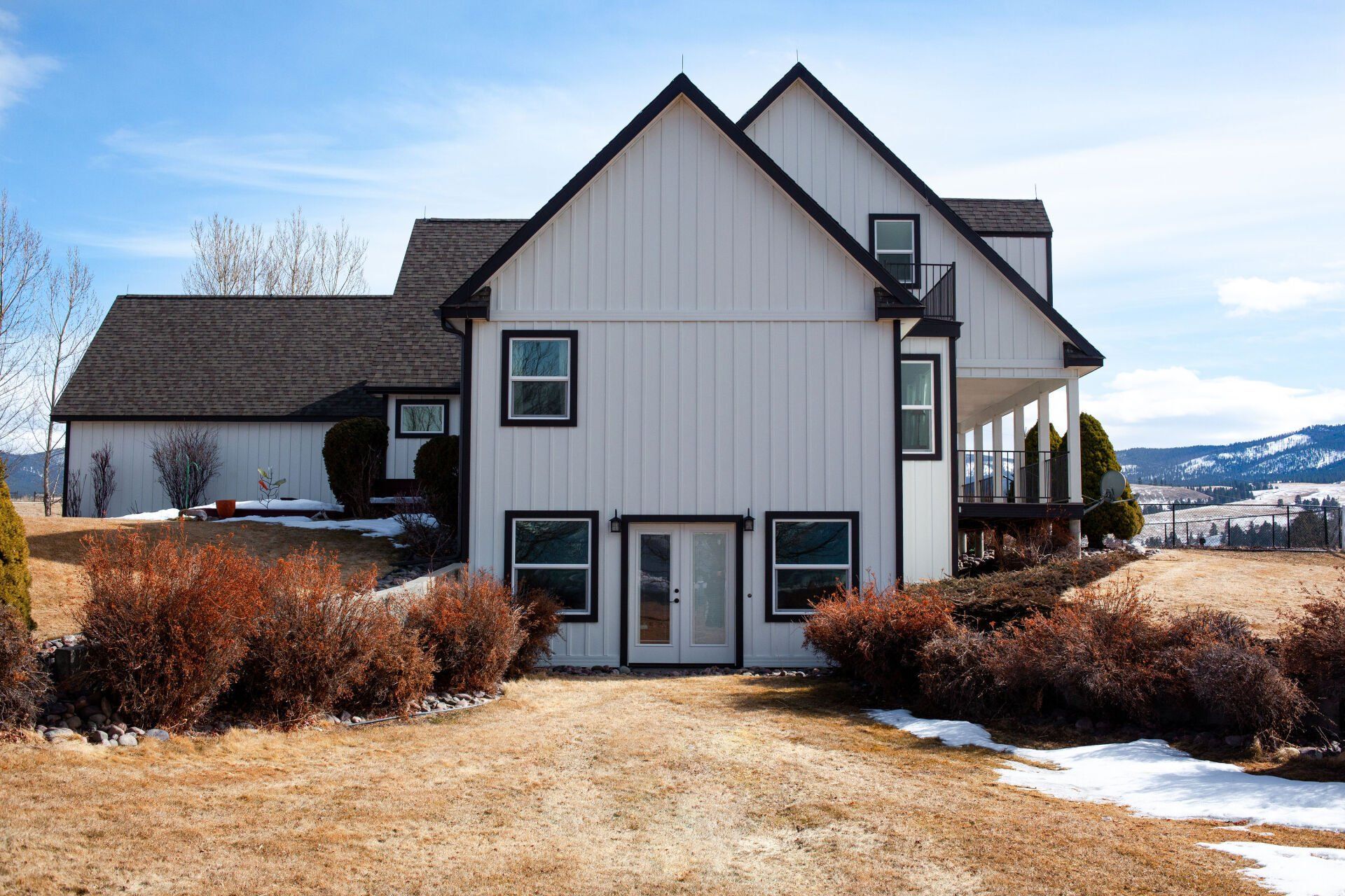 White farmhouse with black trim and a dark roof, brown driveway, and wintery landscape.