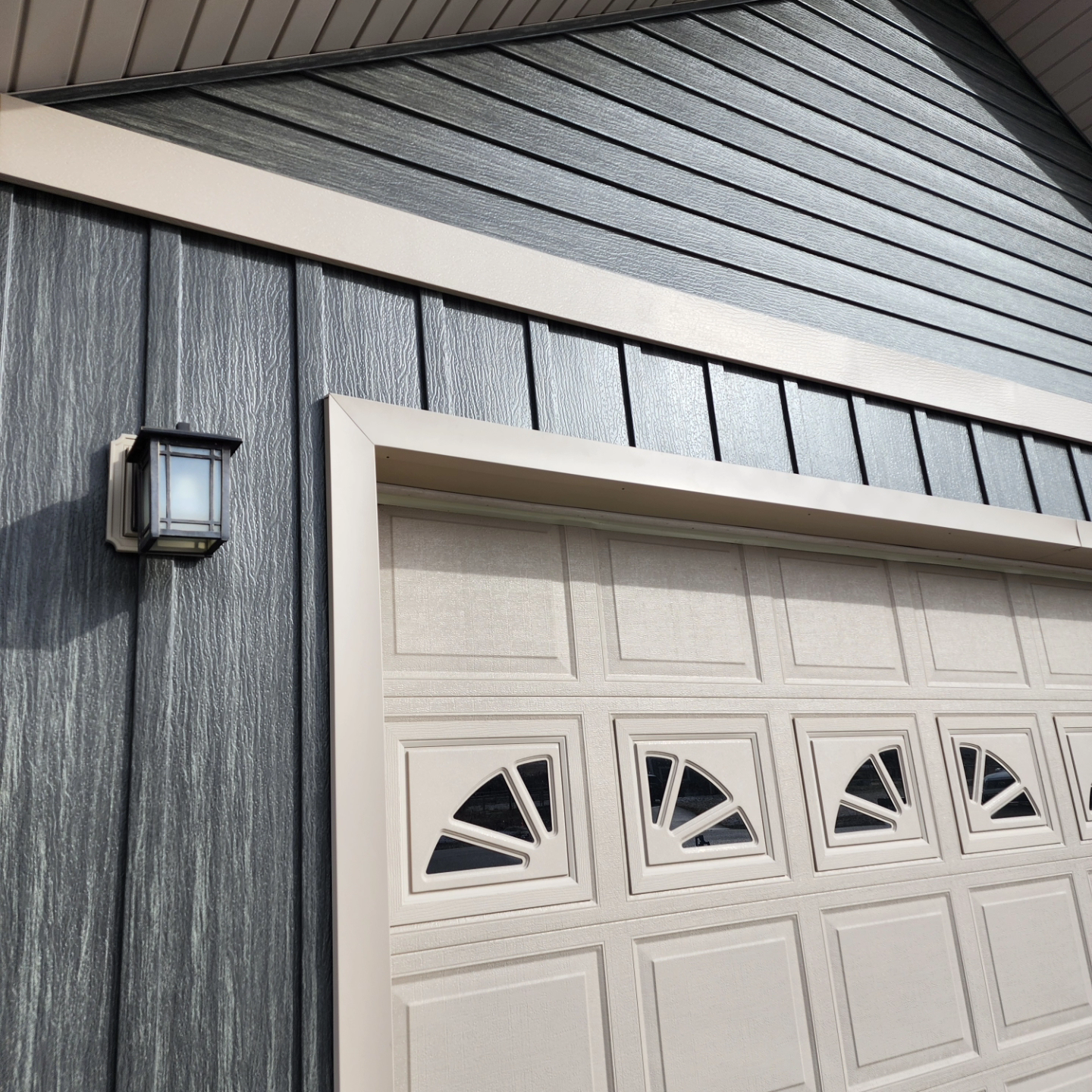 Garage exterior with dark gray siding, tan garage door, and a wall-mounted light.