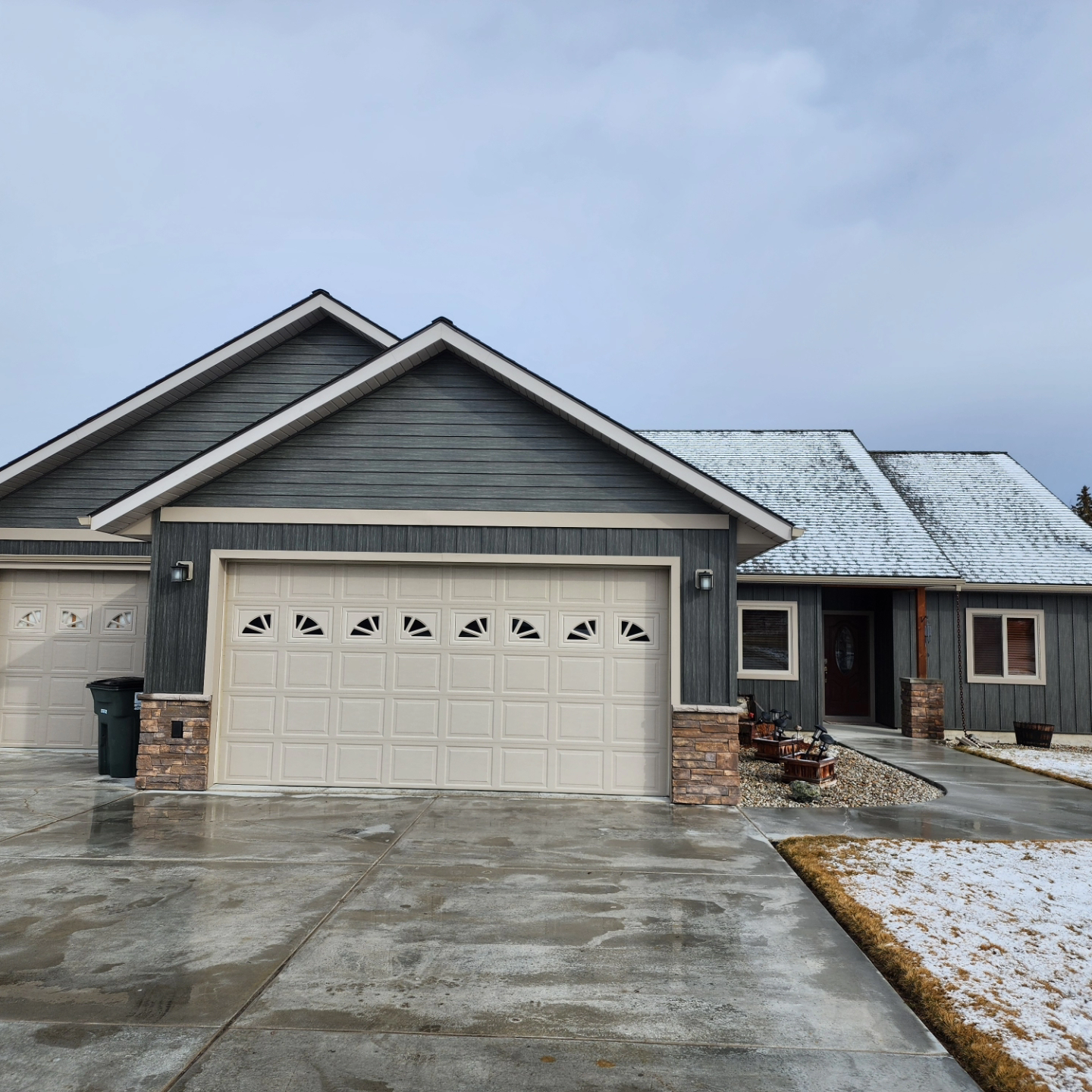 Gray house with snow-covered roof, light beige garage doors, and brick accents on the exterior.